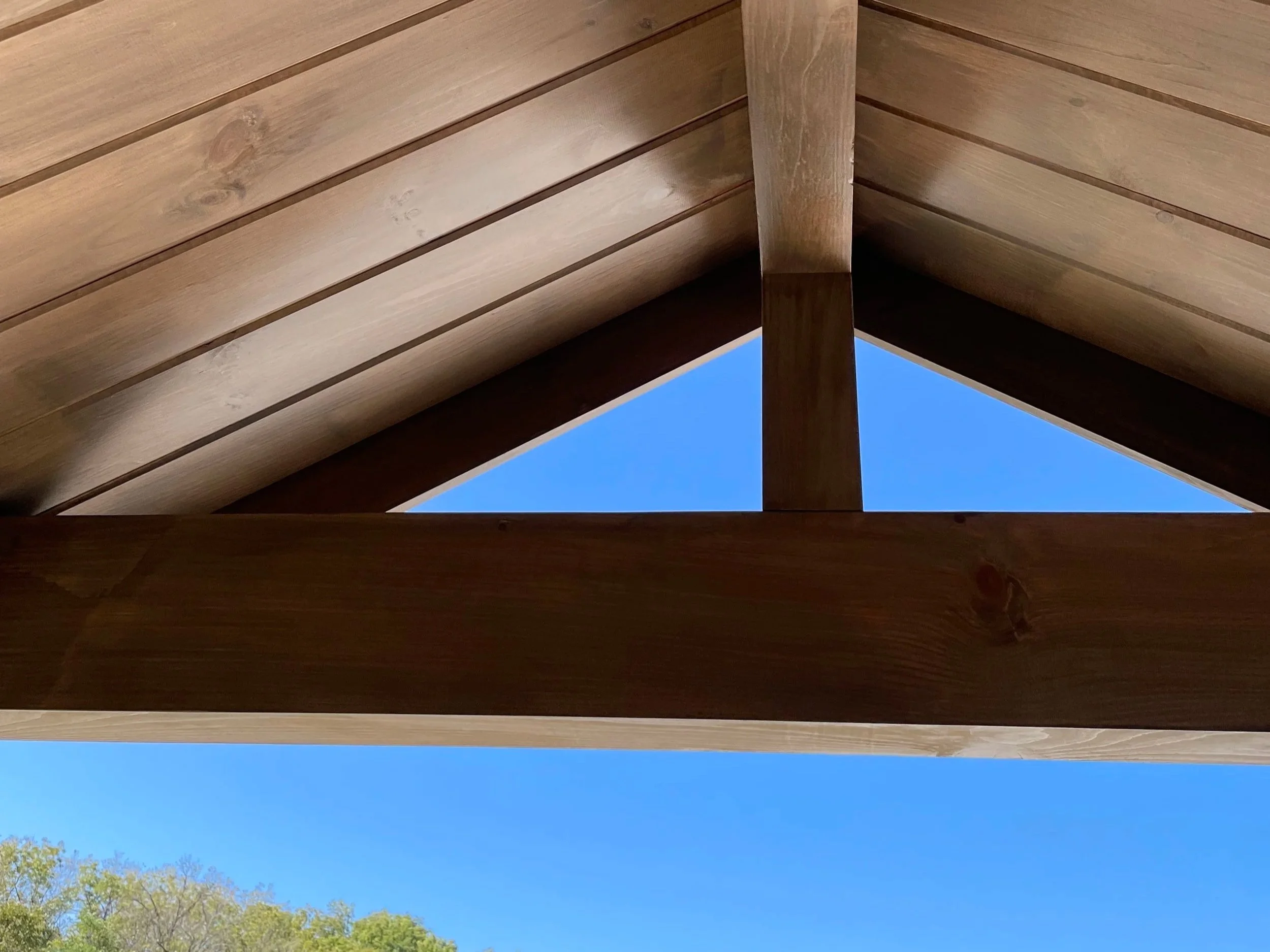 Close-up view of a wooden roof structure with visible wood grain and a clear blue sky in the background. The image appears to be taken from below the roof, showing the underside of the wooden beams and ceiling.