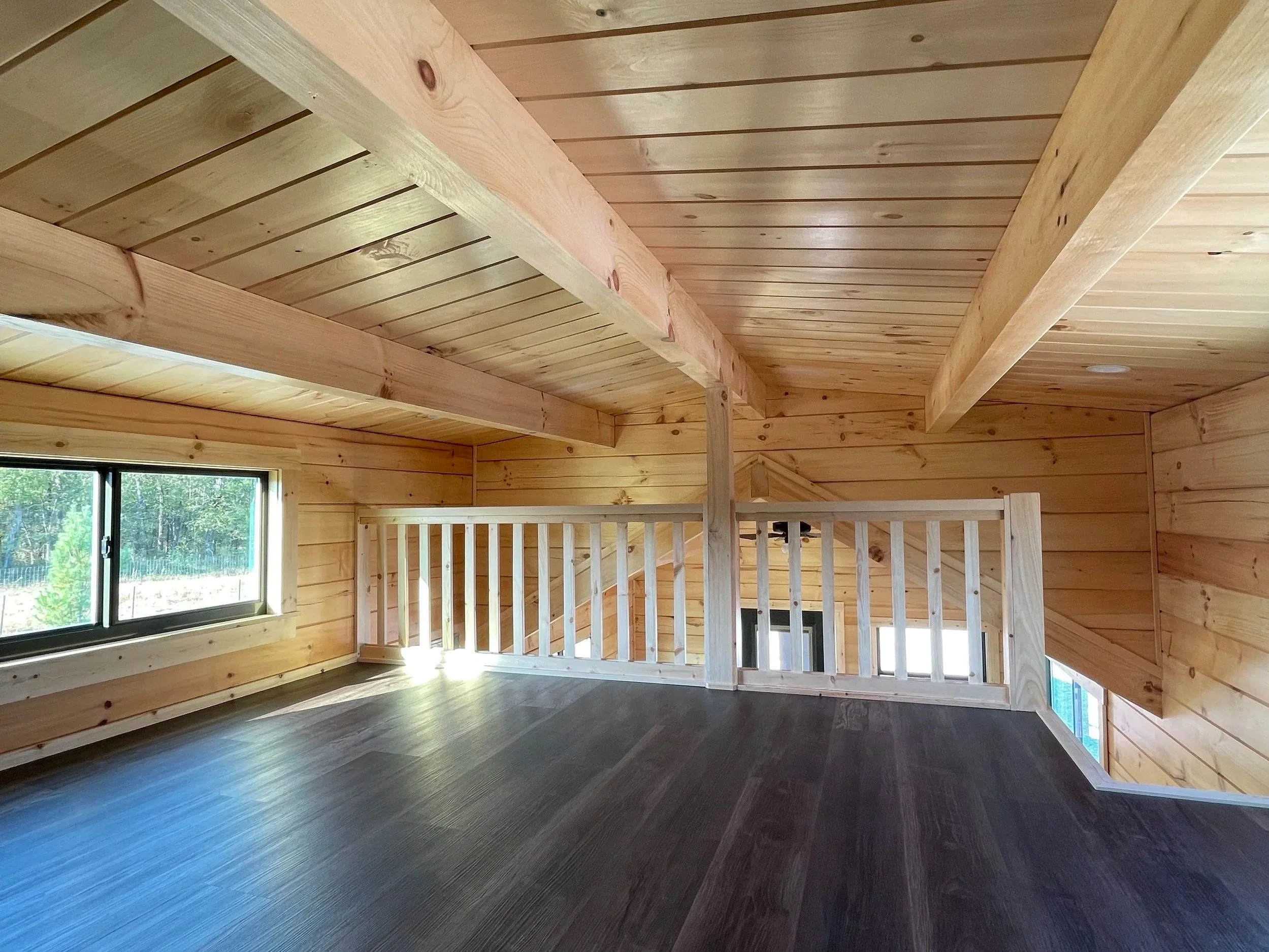 Interior of a wooden house with a vaulted ceiling and a large window, showing trees outside. The room has wood paneling on the walls and dark flooring.