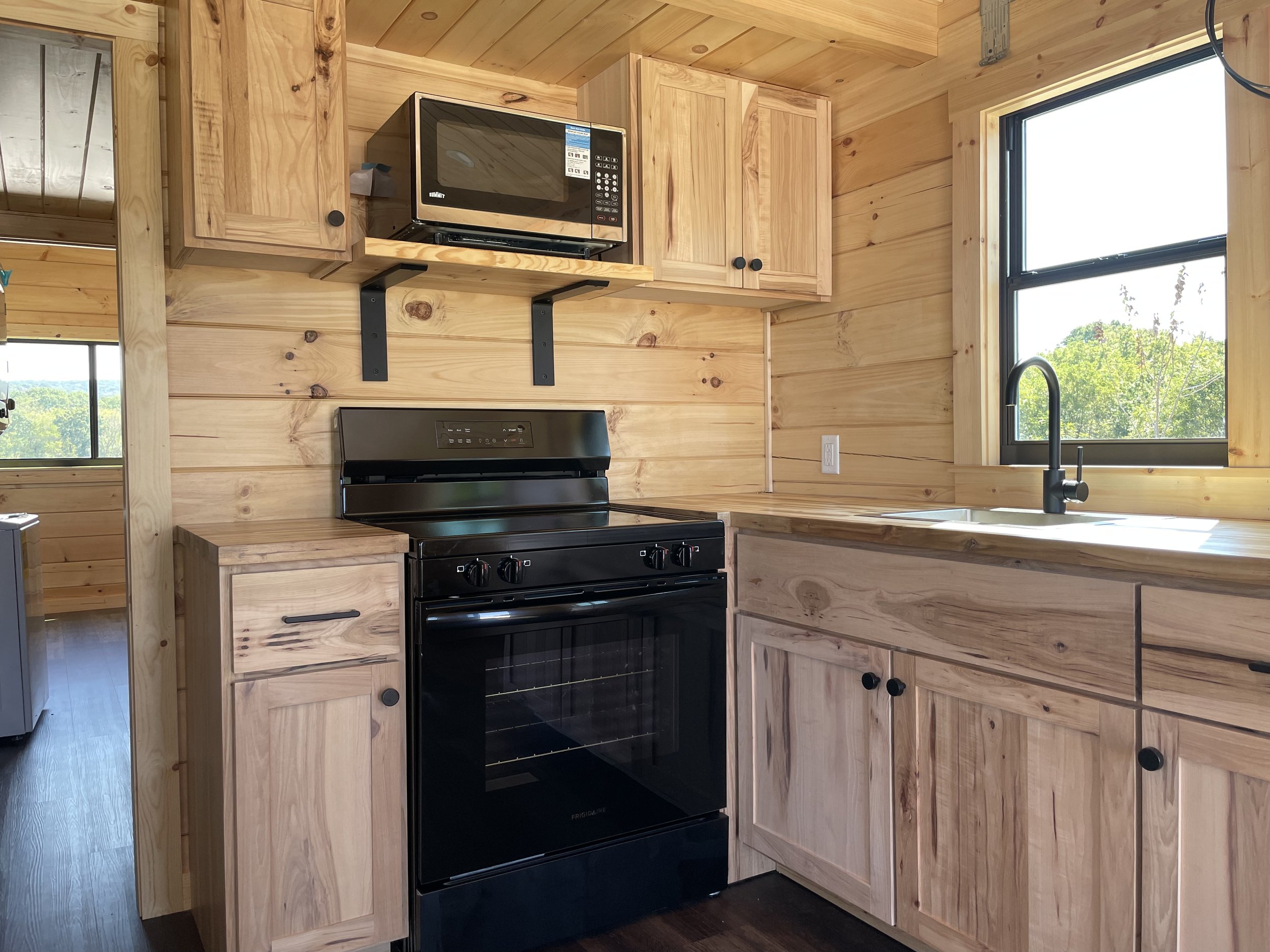 Kitchen with wood-paneled walls, cabinets, and ceiling. Black oven and microwave, window above the sink with a black faucet, and natural light.