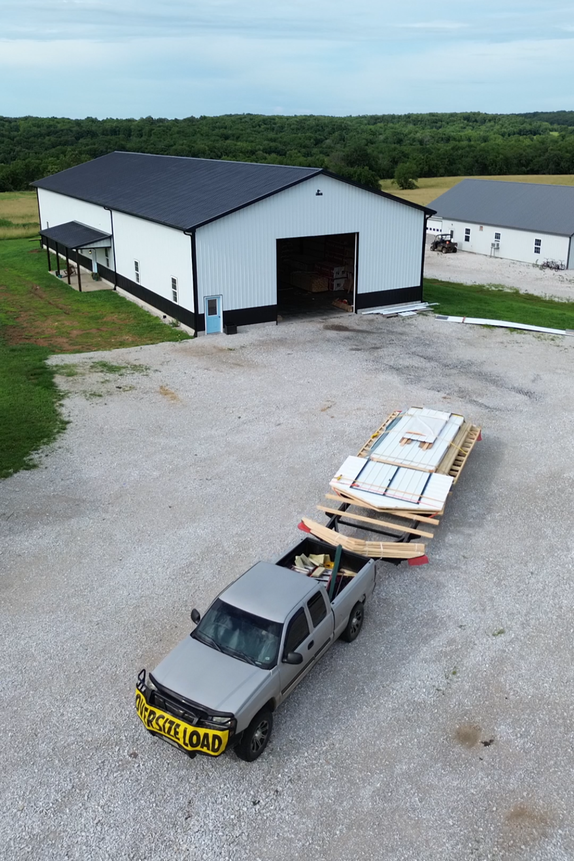 A gravel lot with a silver pickup truck loaded with construction materials, pulling a trailer with more building supplies, parked in front of a large white and black barn under a cloudy sky.