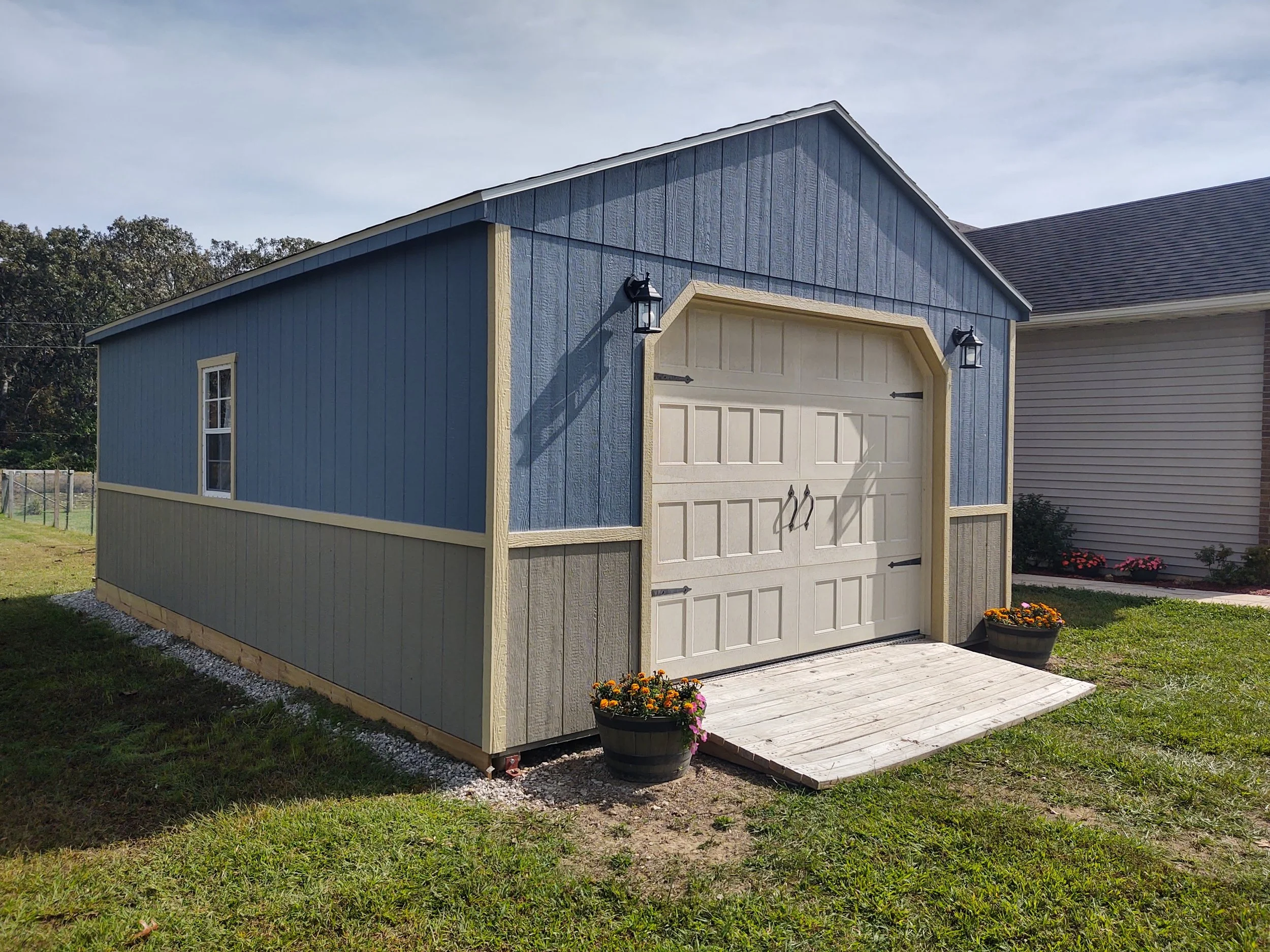 A blue and beige detached garage with a white garage door, mounted black outdoor lanterns, a small wooden deck in front, and potted flowers on each side, surrounded by a grassy yard with trees and neighboring houses in the background.