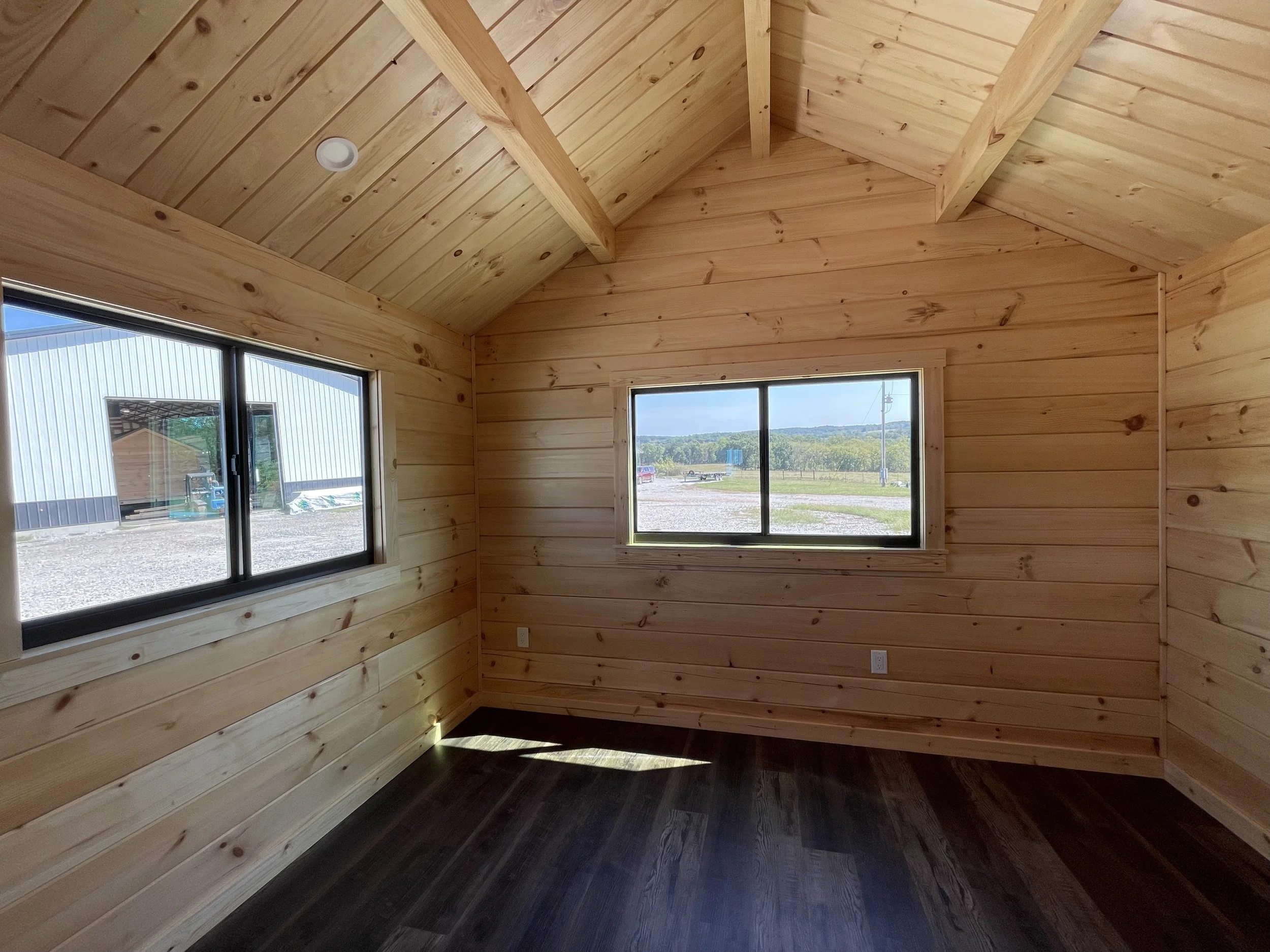 An interior view of a wooden room with natural wood paneling and two large windows showing an outdoor scene of a rural landscape with trees, a gravel driveway, and a building outside.
