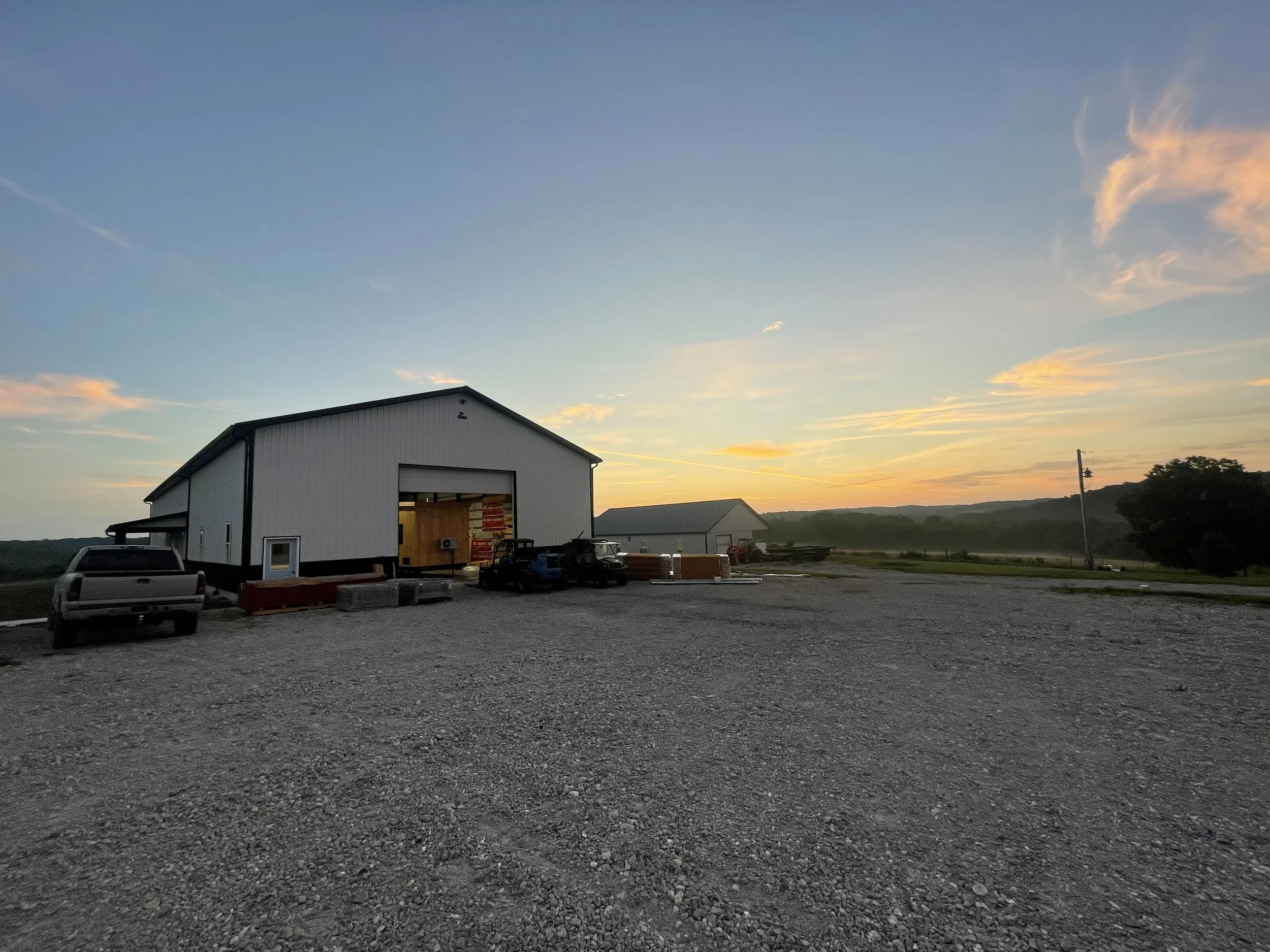 A large white barn with an open door at sunset, with gravel parking area and vehicles nearby, in a rural setting with open fields and trees.