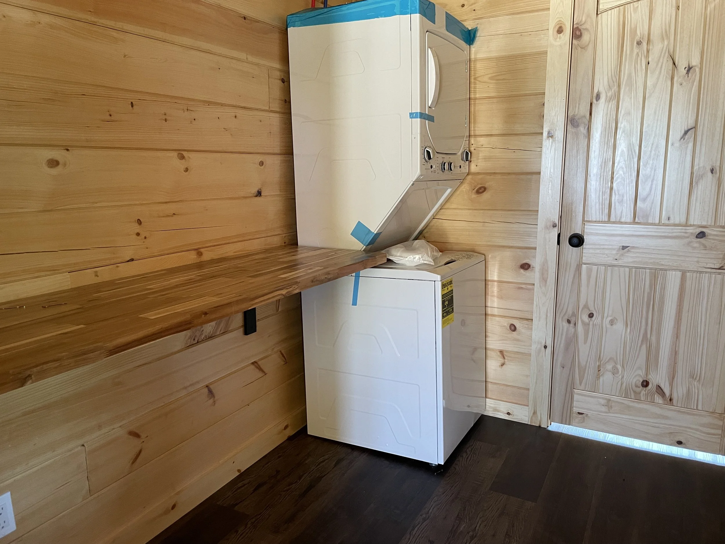 A white top-loading washing machine is placed against a wooden wall and ceiling, with blue painter's tape at the top corner, and a white plastic bag on top.