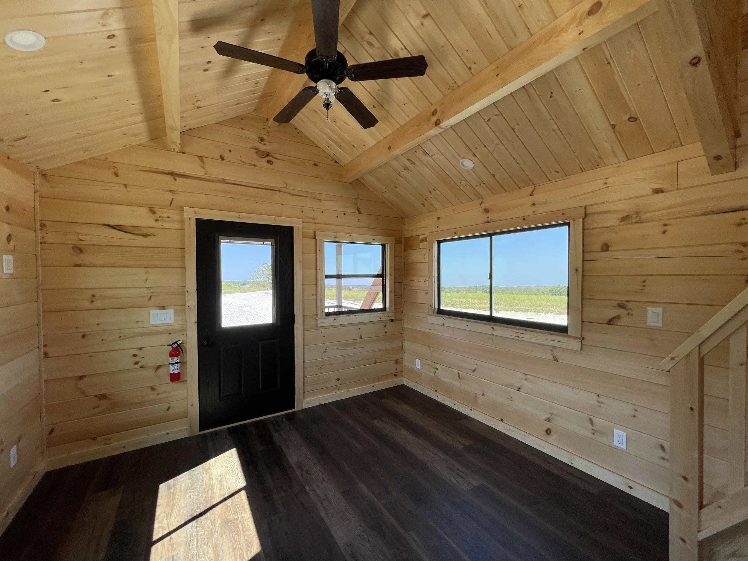 Interior of a wooden room with a black ceiling fan, large windows showing outdoor scenery, and a black door.