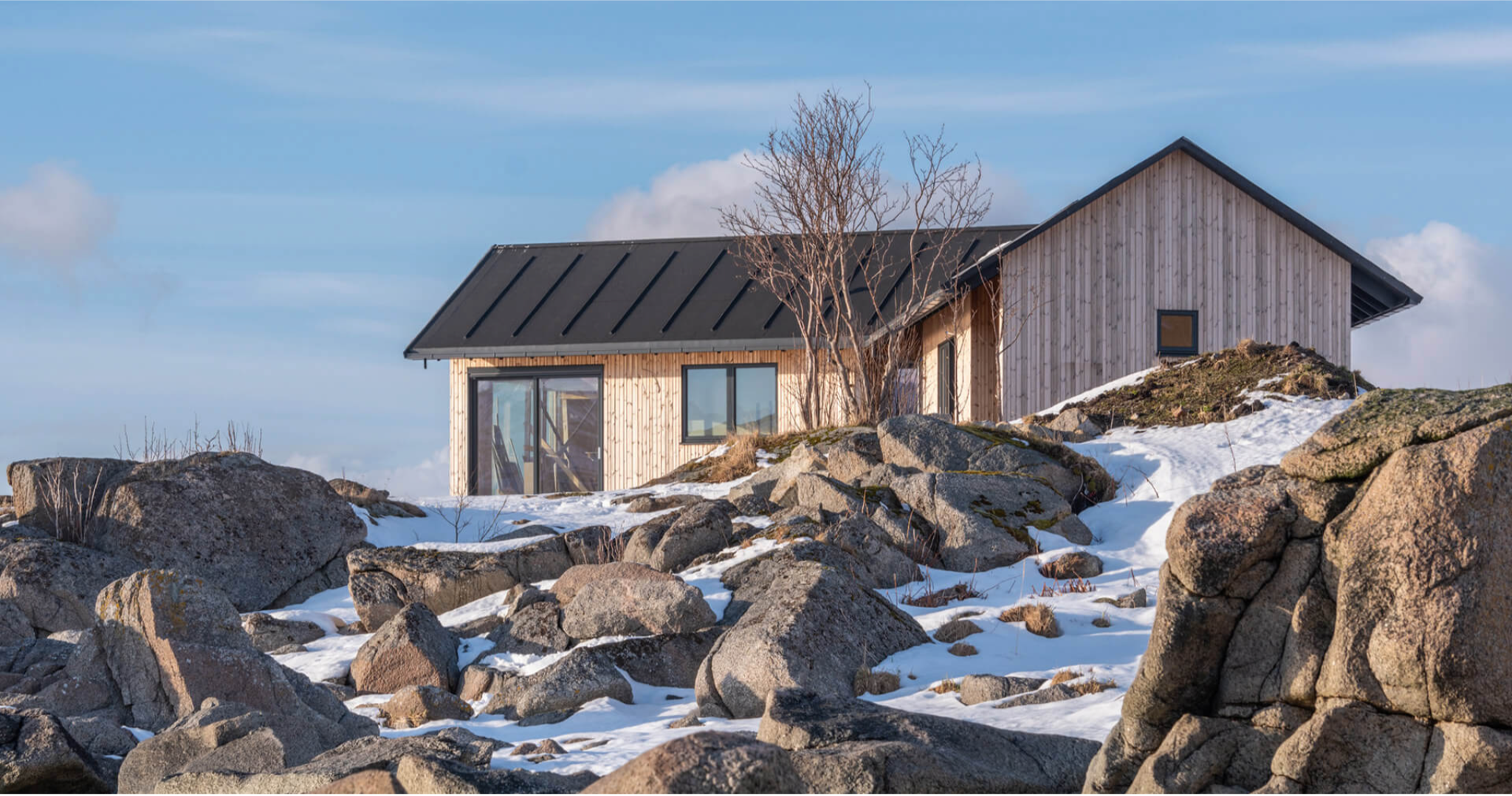 A modern house with wooden Lunawood Thermowood  exterior and black metal roof situated on a snowy rocky hillside, with a barren tree near the house and a blue sky with clouds in the background.