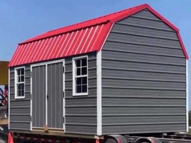 Gray shed with white trim and a red metal roof on a trailer. Built by Premium Structures. Transported by D&S Transports