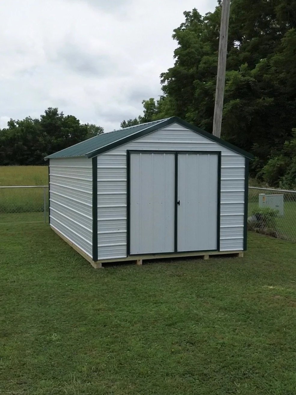 Small white metal storage shed with a green roof sitting on wooden skids in a grassy yard, surrounded by a chain-link fence and trees. Built by Premium Structures LLC.