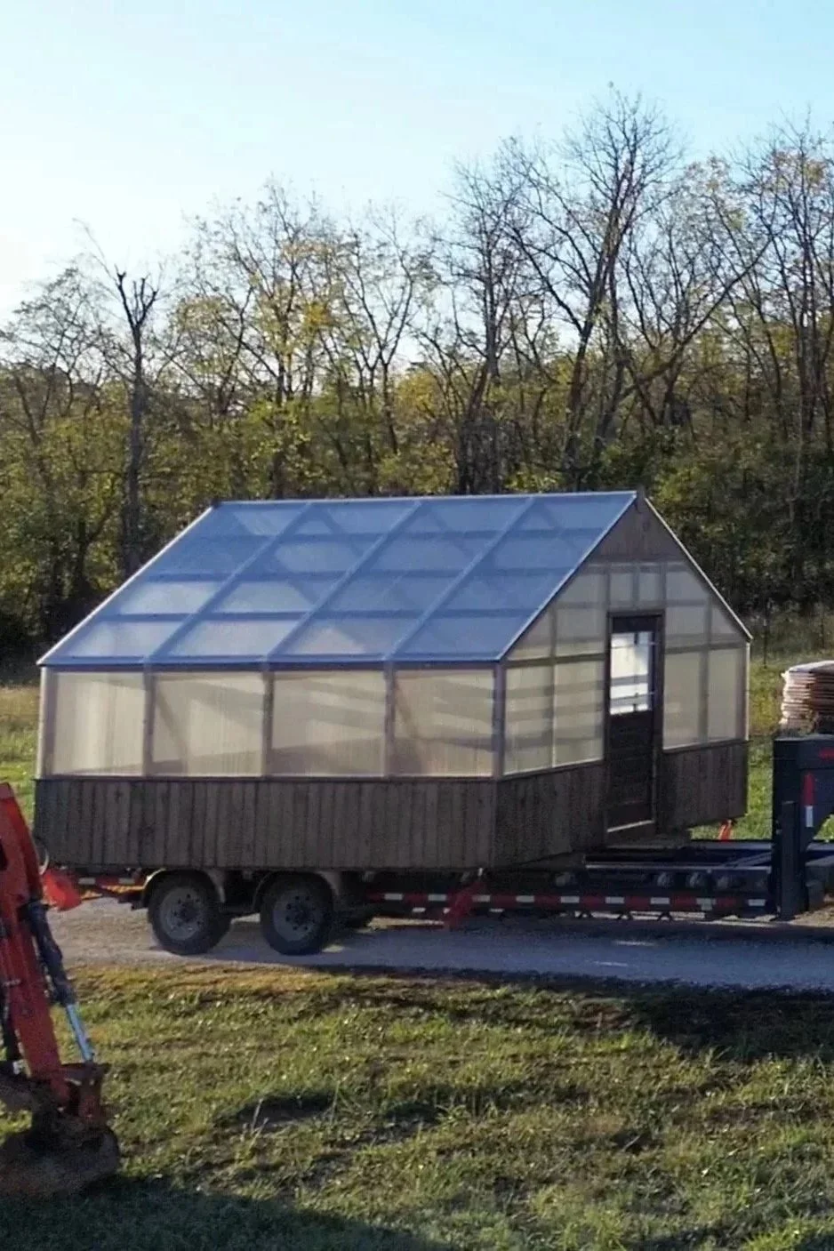 Small greenhouse with a transparent roof and walls on a trailer, set outdoors with trees in the background.