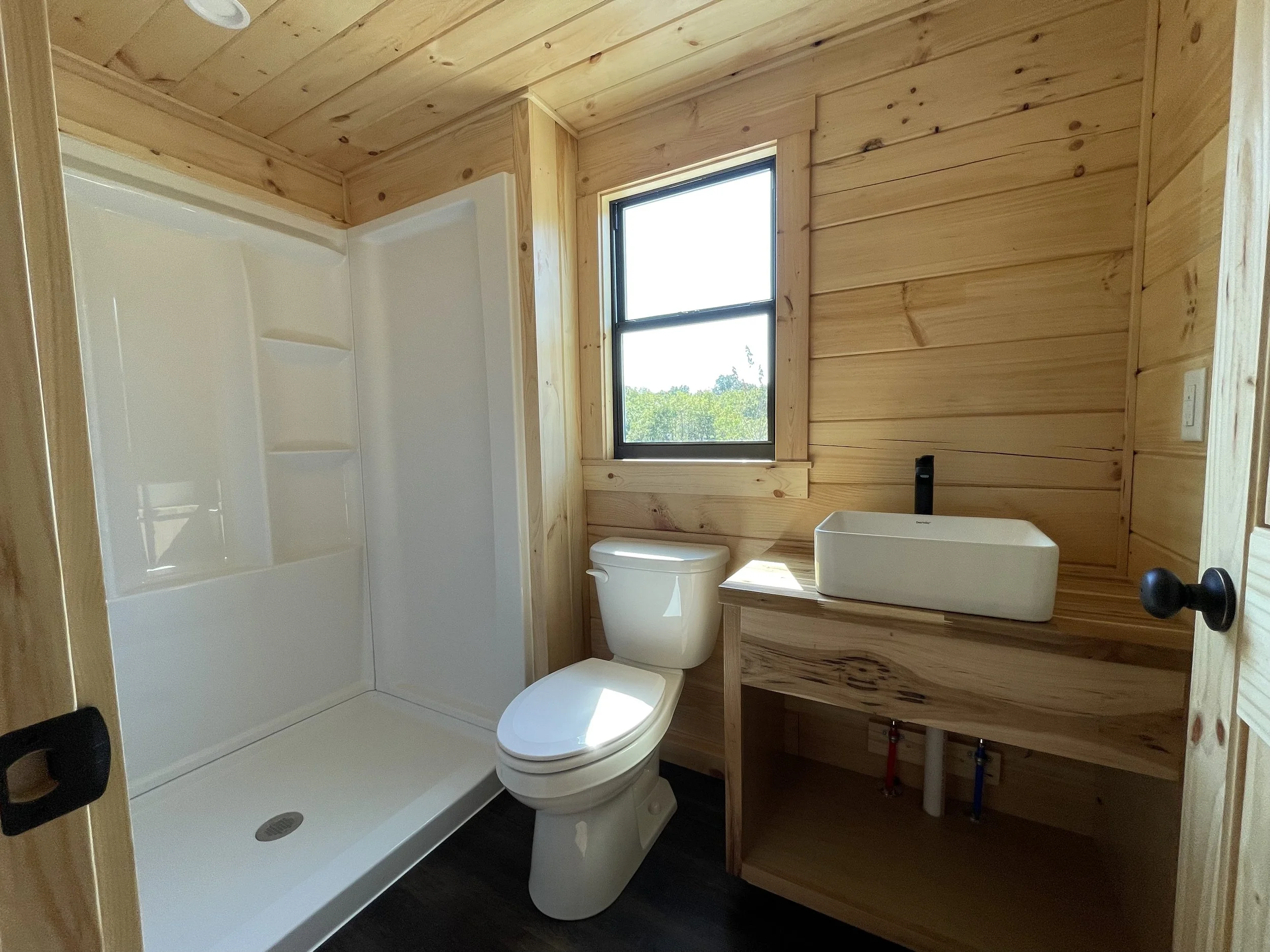 A bathroom with wooden paneling on the walls and ceiling. It has a window and a shower stall, a toilet, a rectangular sink, and a black handle. There are visible plumbing pipes under the sink.