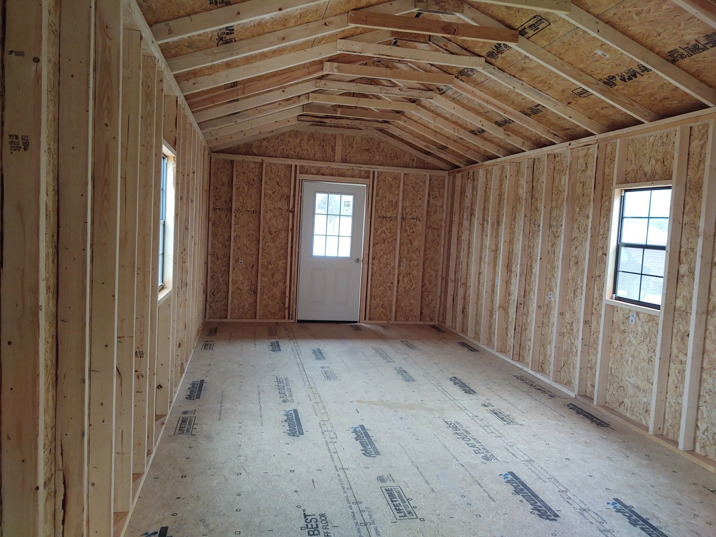 The interior of an unfinished room in a house under construction, with exposed wooden framing, wall studs, ceiling joists, and two windows.