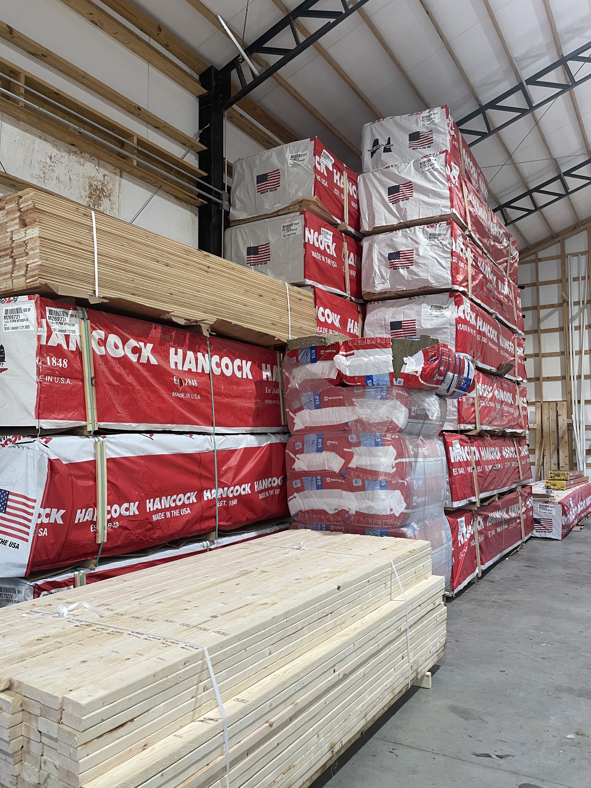 Construction materials in a warehouse, including stacked wooden planks and packages of drywall with red and white packaging labeled 'Hancock.'