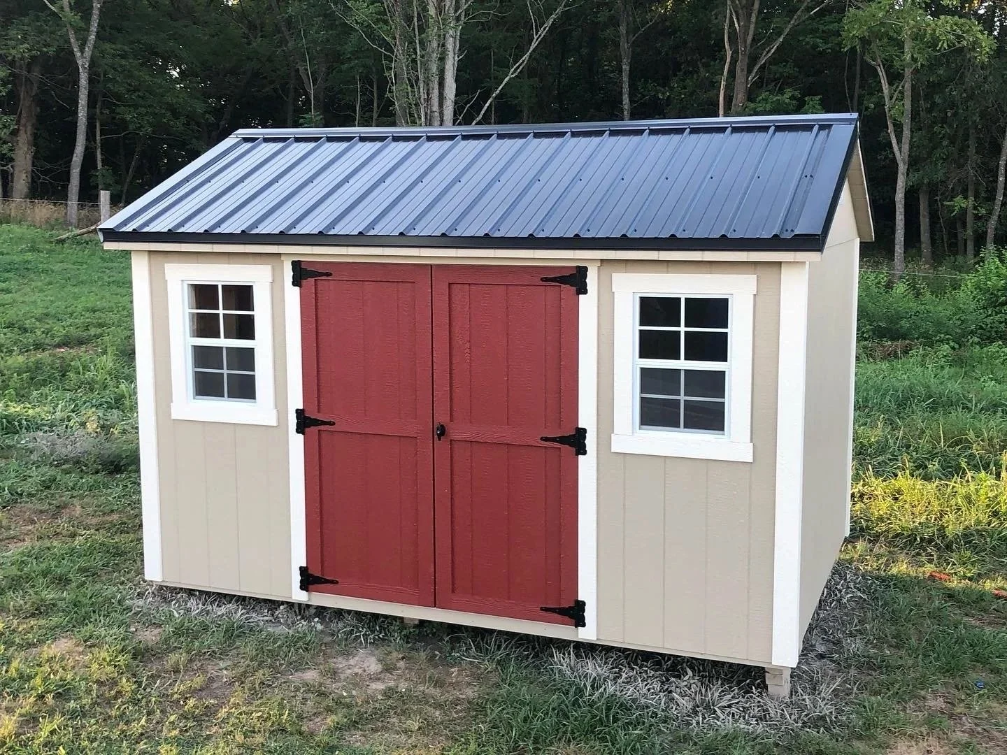 A small outdoor shed with beige walls, white trim, black hinges, a red double door, and two small windows. The shed has a metal roof and is located on a grassy area with trees in the background. Built by Premium Structures LLC.