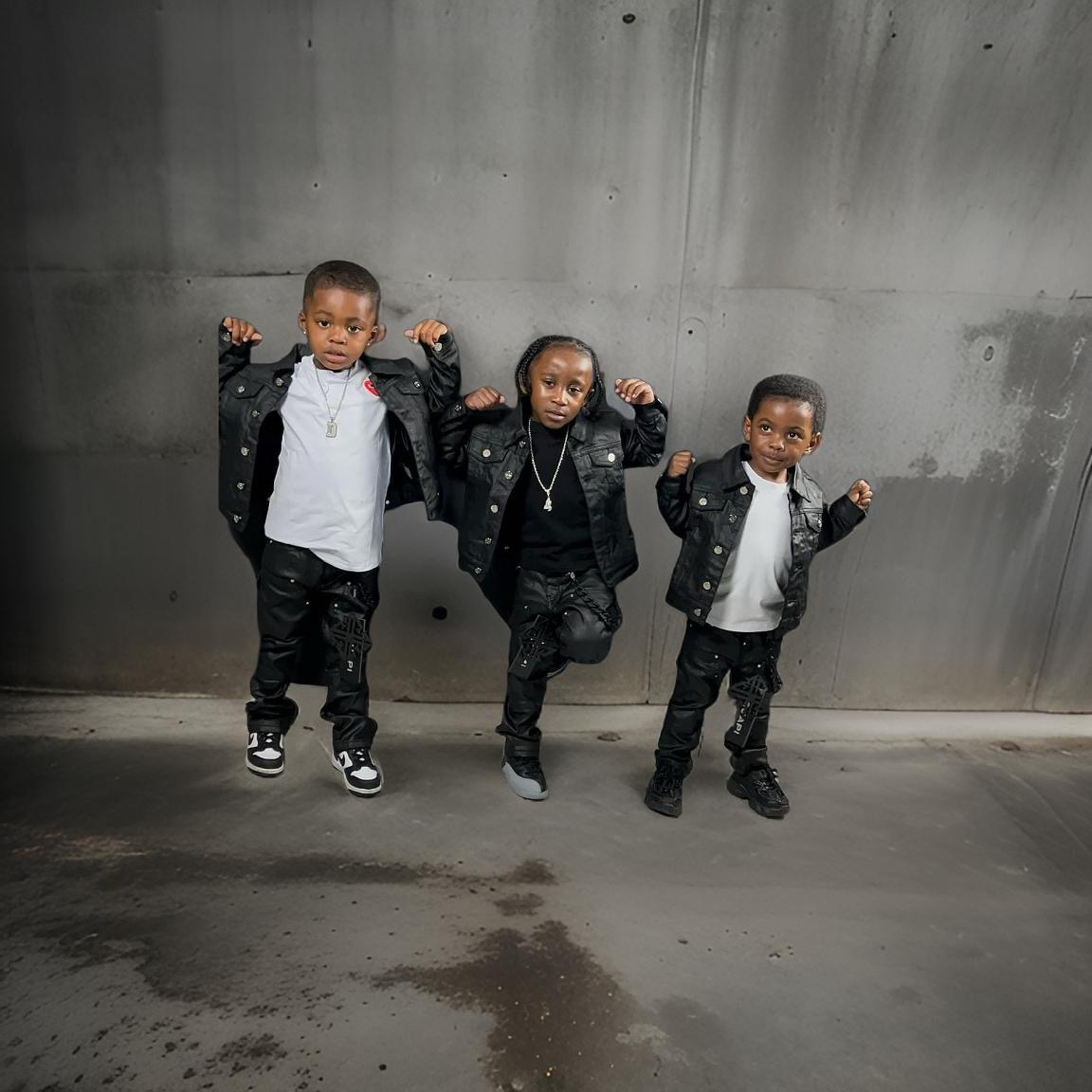 Three children wearing black jackets and black pants, with confident poses, standing in front of a gray concrete wall.