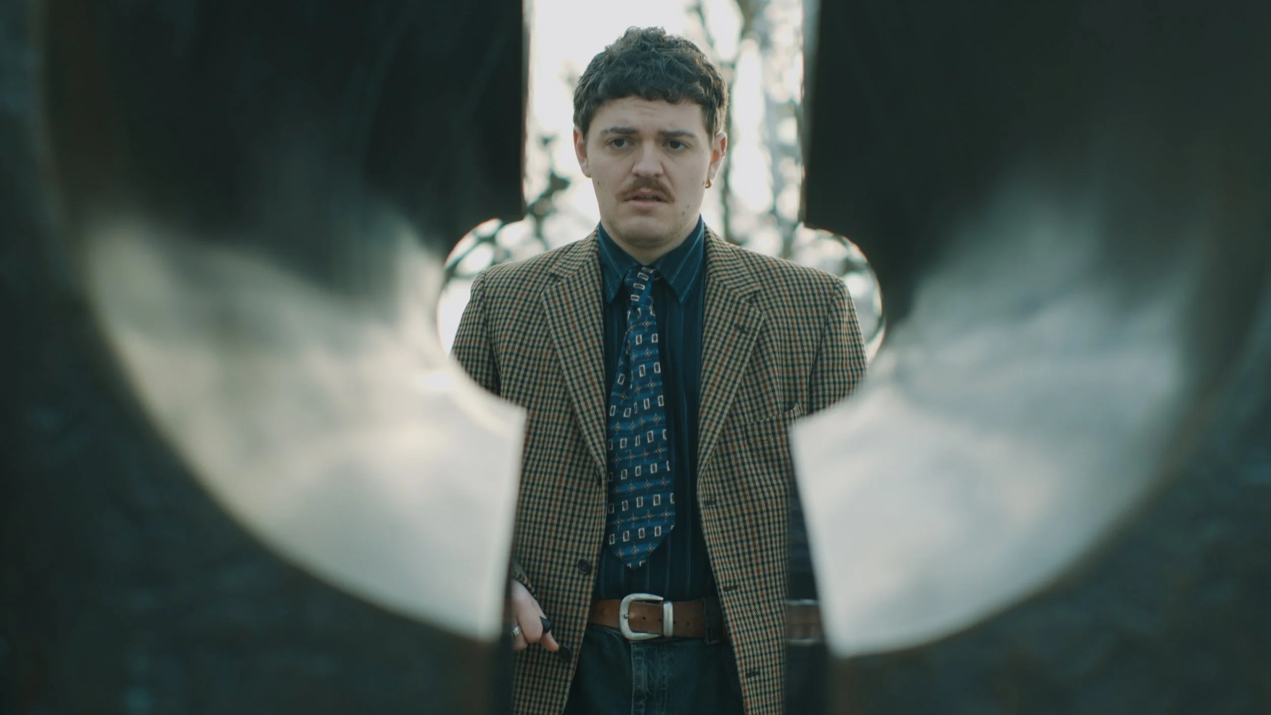 Man wearing a plaid blazer and patterned tie looking at the camera through a gap in two objects.