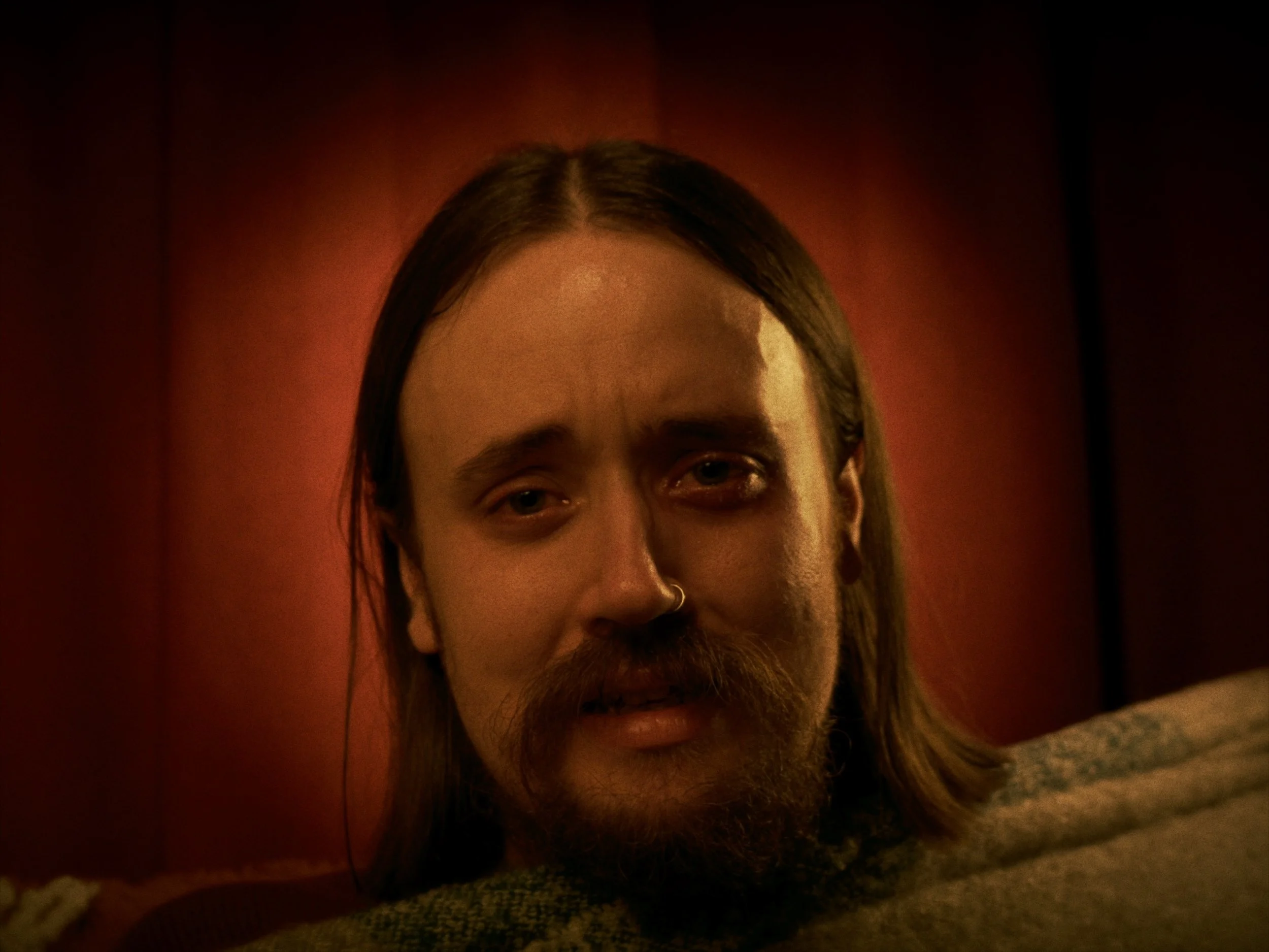 A man with long hair, a mustache, and a nose ring lying on a bed with a red headboard, looking at the camera with a sad expression.