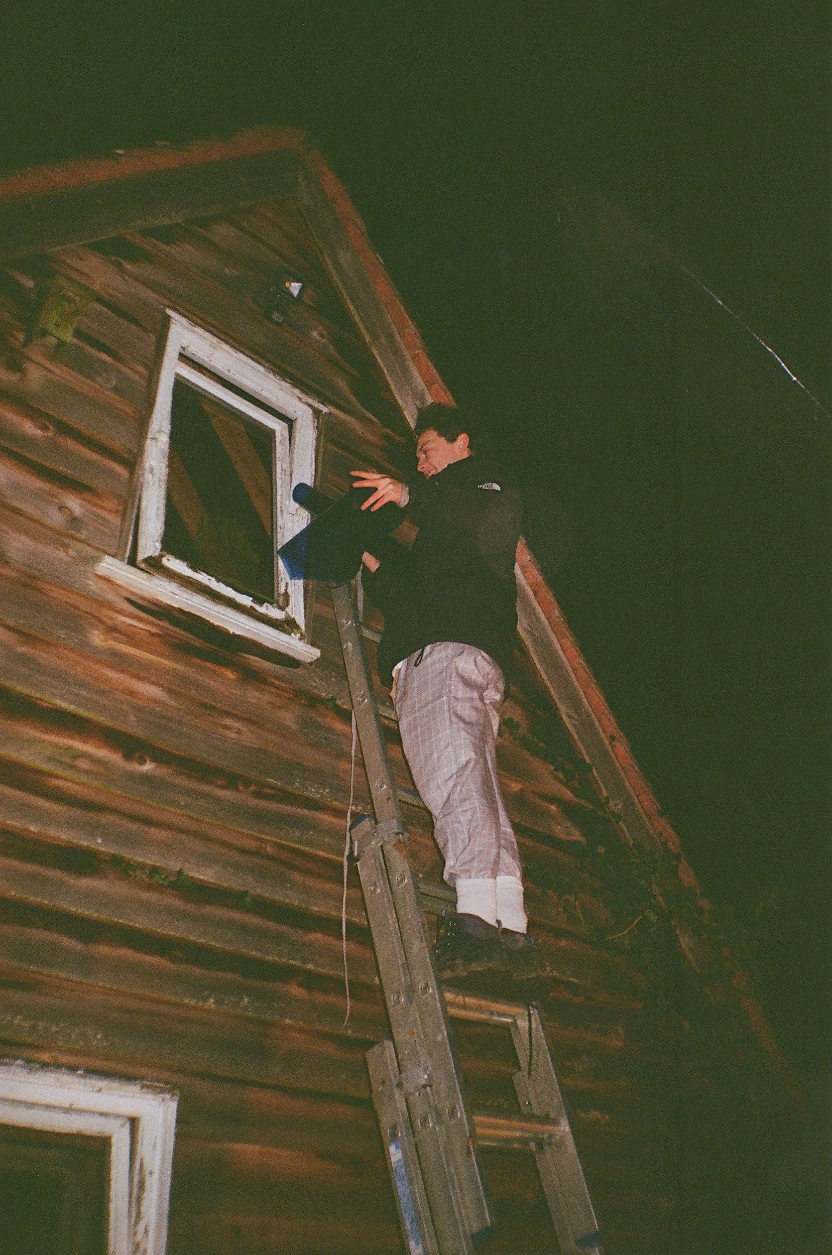 Person standing on a ladder cleaning or looking out of a window on a wooden house at night.