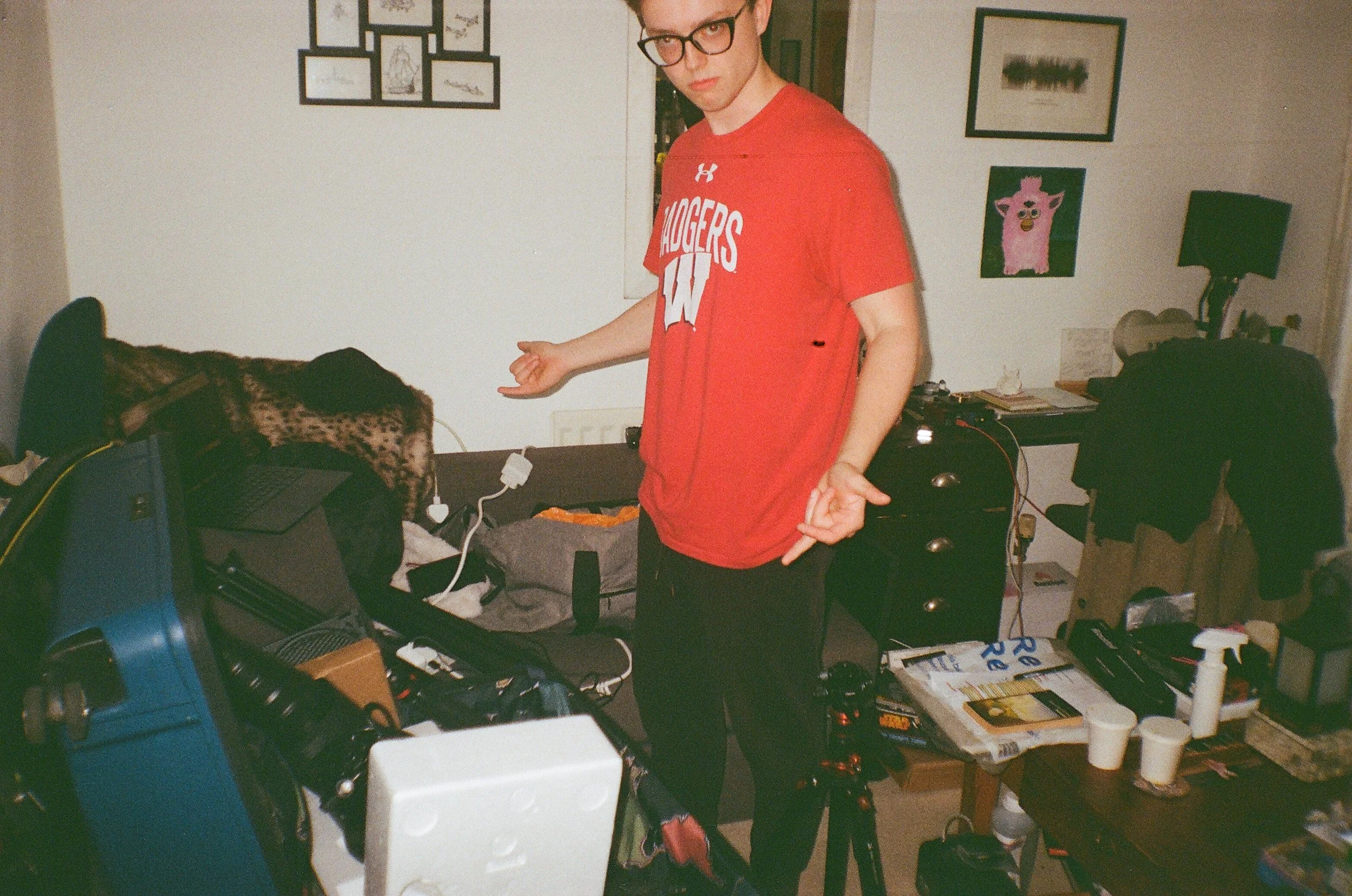 Young man in red t-shirt and glasses gesturing in a cluttered living room with furniture, artwork, and electronic devices.