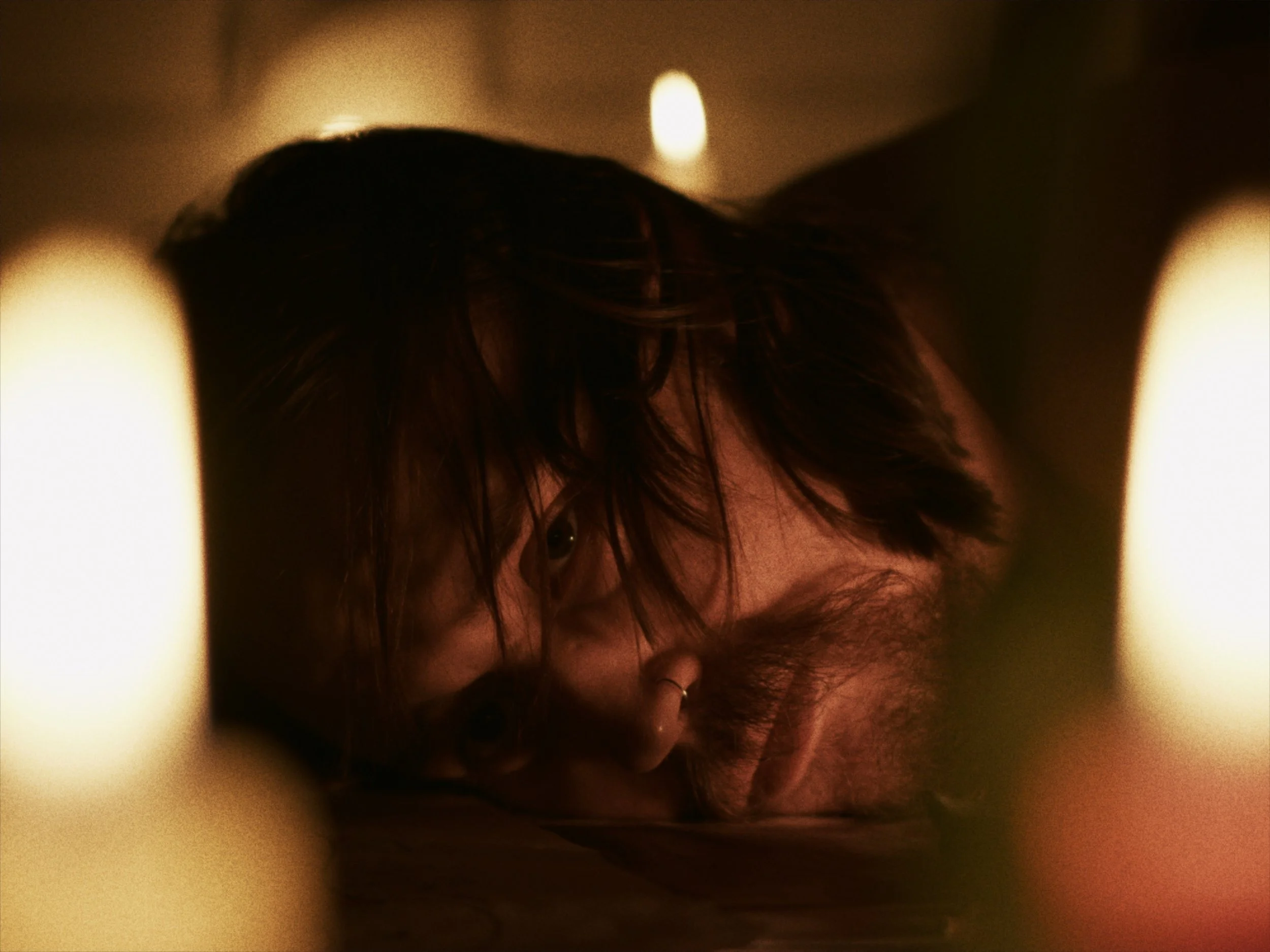 A person with dark hair and glasses lying down in a dimly lit room, surrounded by small candles.