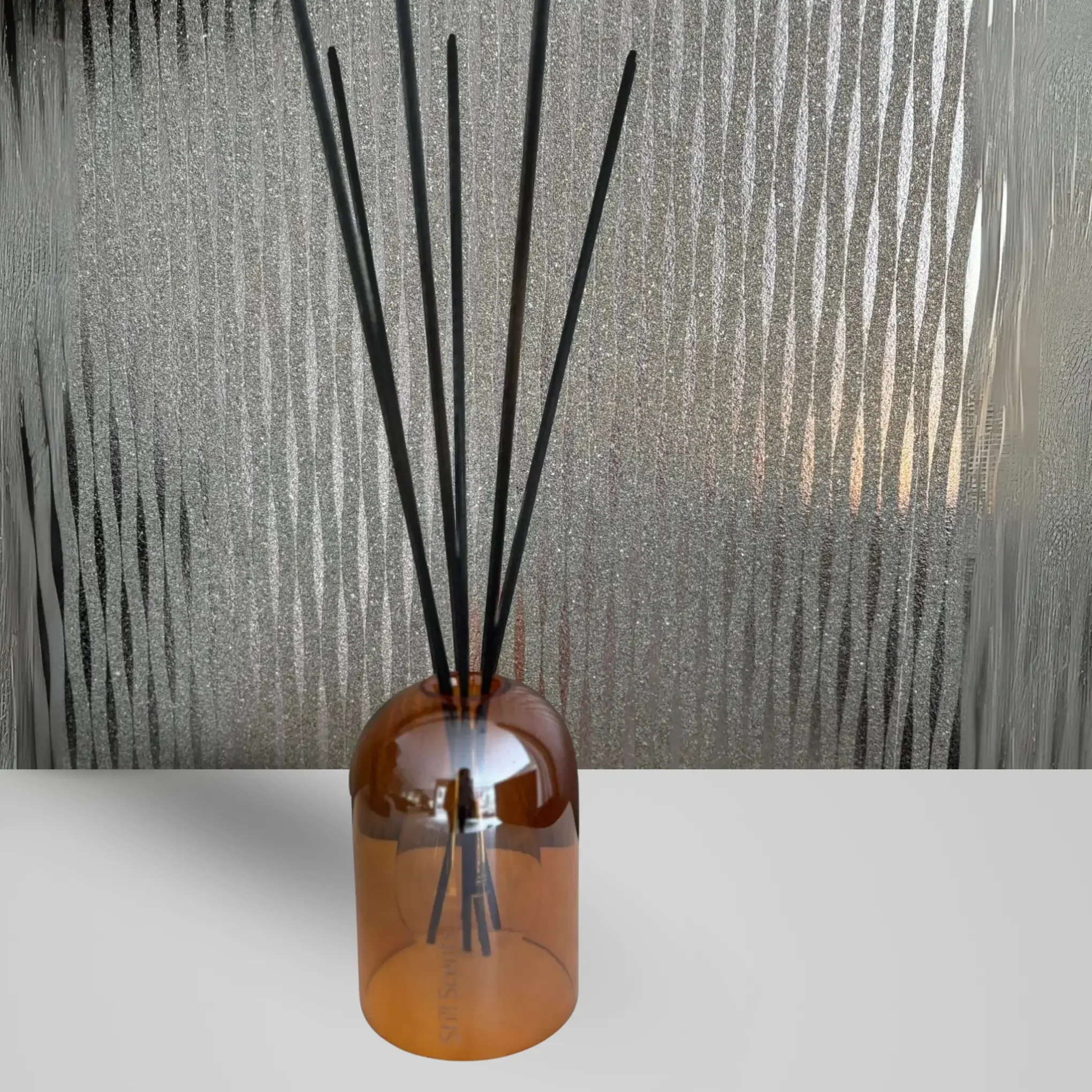 Brown glass diffuser bottle with black reed sticks on a white surface, textured gray wall in the background.