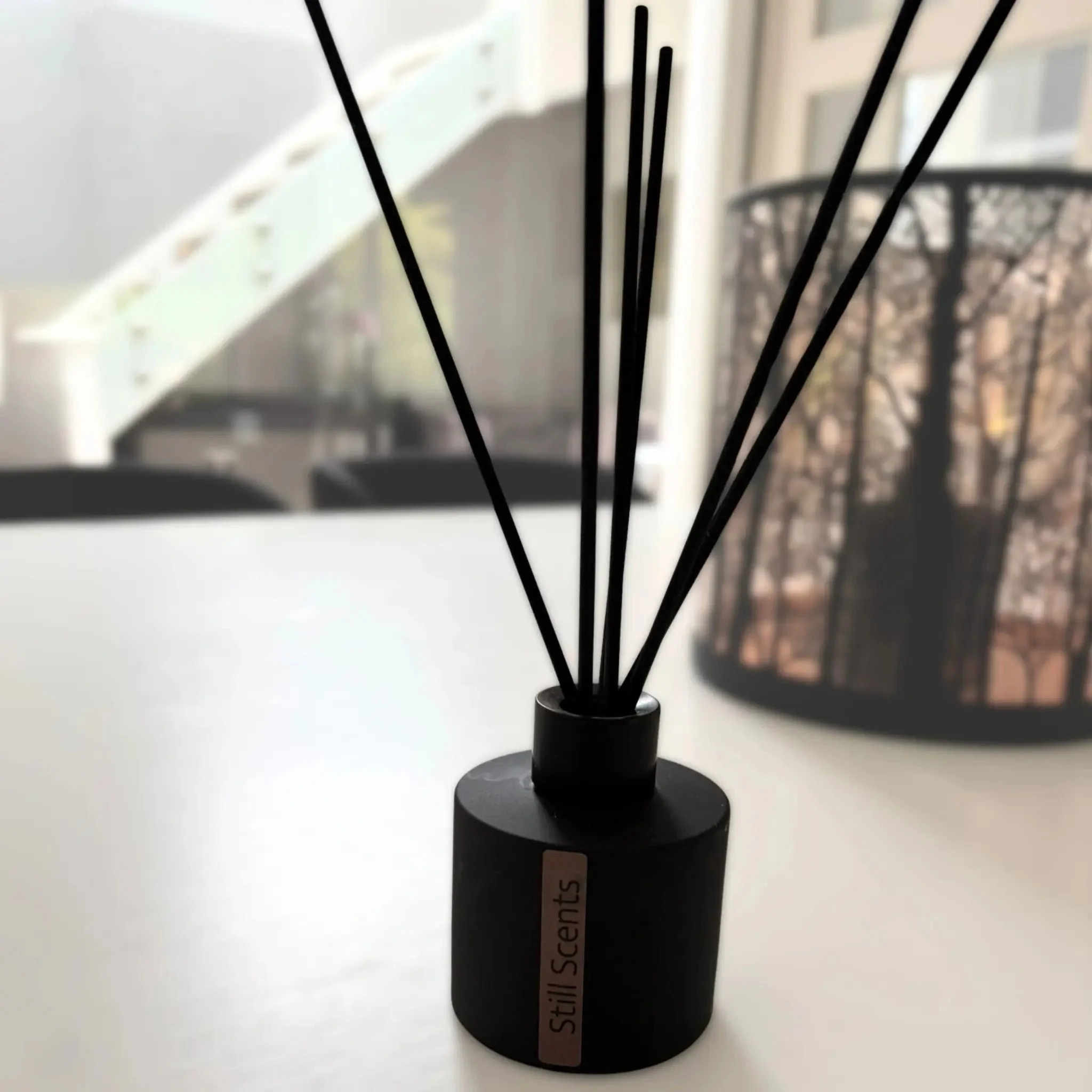 A black reed diffuser with a label that says "Still Scents" on a white surface, with a blurred background of a balcony and black wire basket.