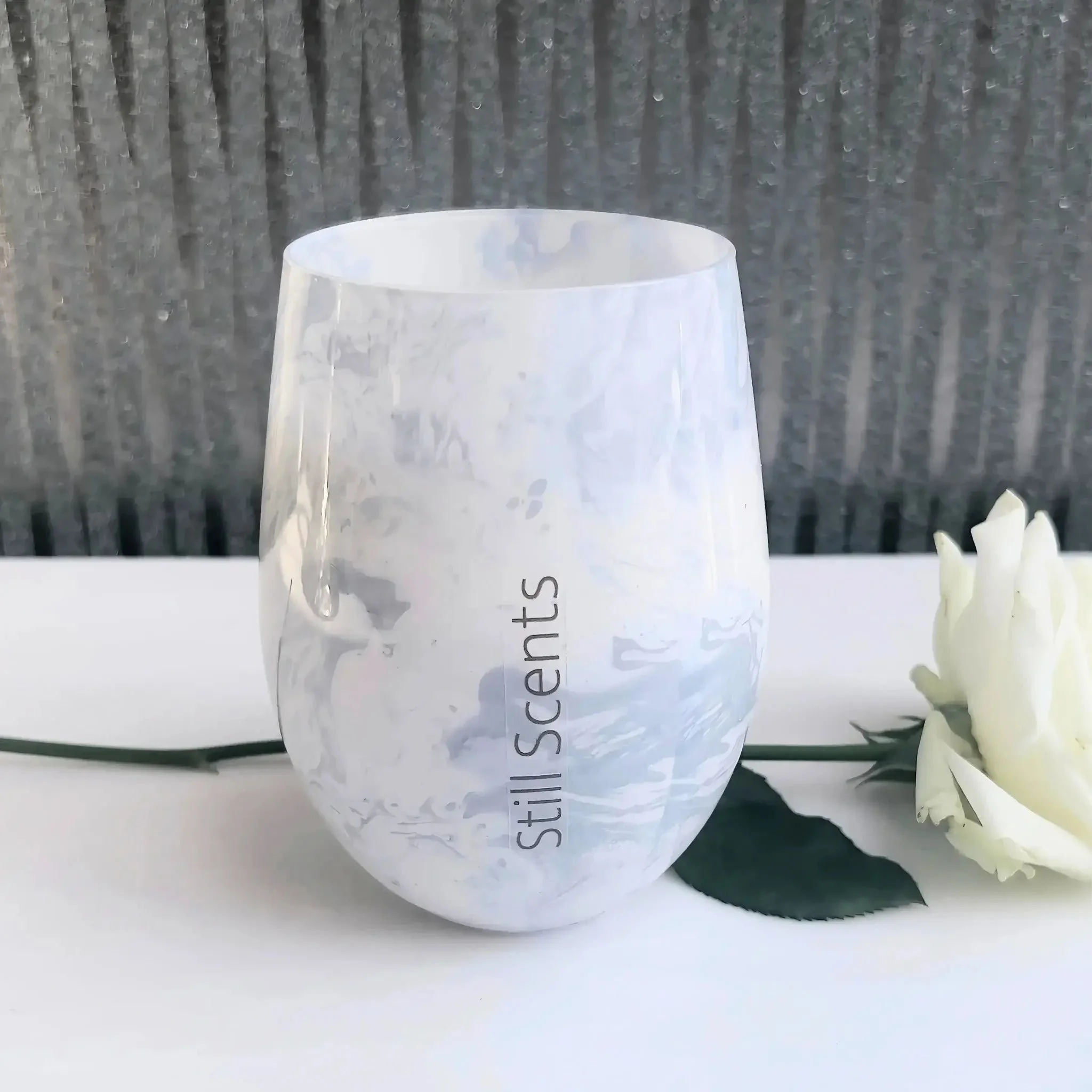 White marbled glass candle with a single cream rose ling behind and shiny wallpaper in the background.