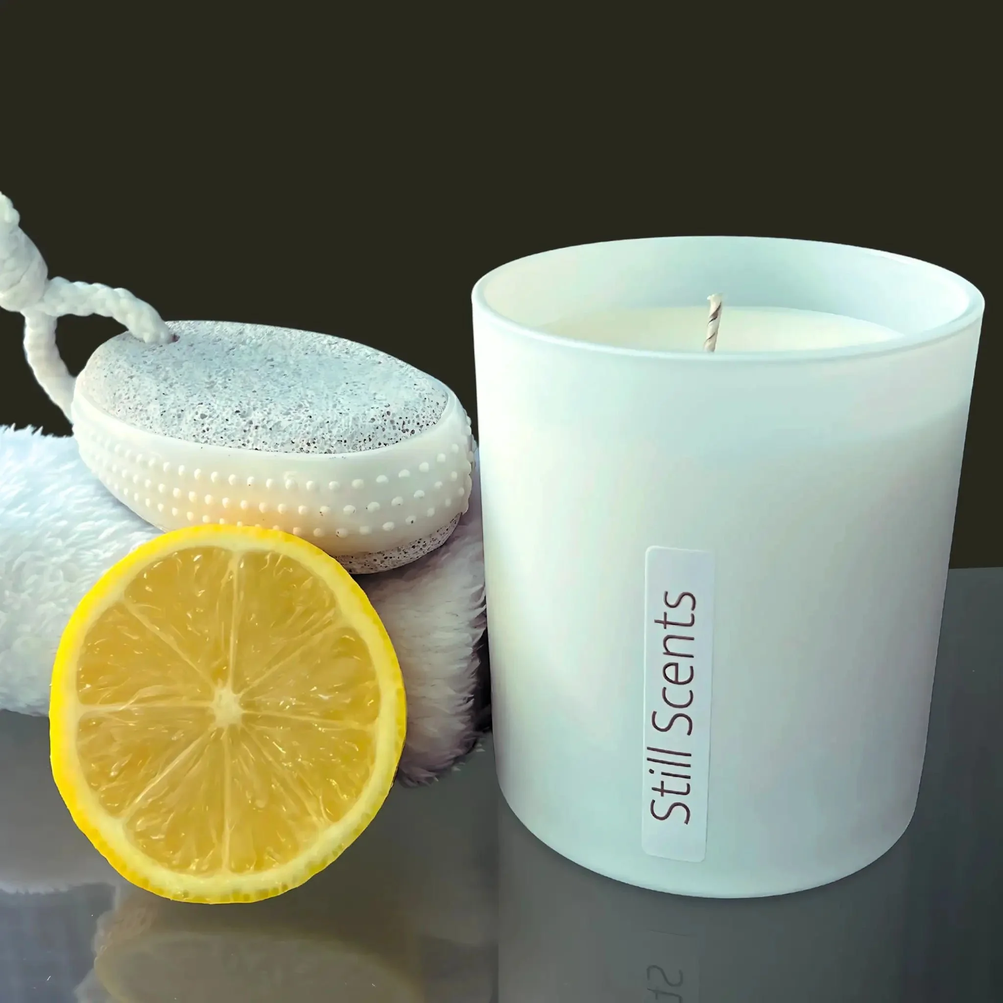 A white candle with a label that reads 'Still Scents', a halved lemon, a white bath lily, and a white bath sponge arranged on a reflective surface against a dark background.
