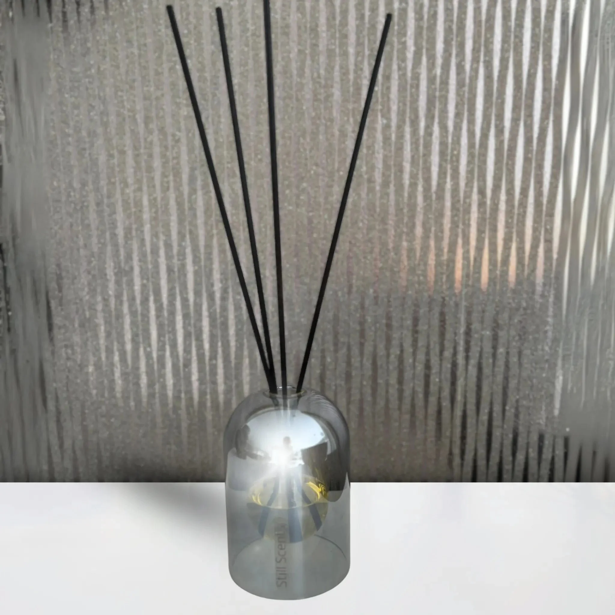 A glass diffuser bottle with black reed sticks on a white surface against a textured glass background.