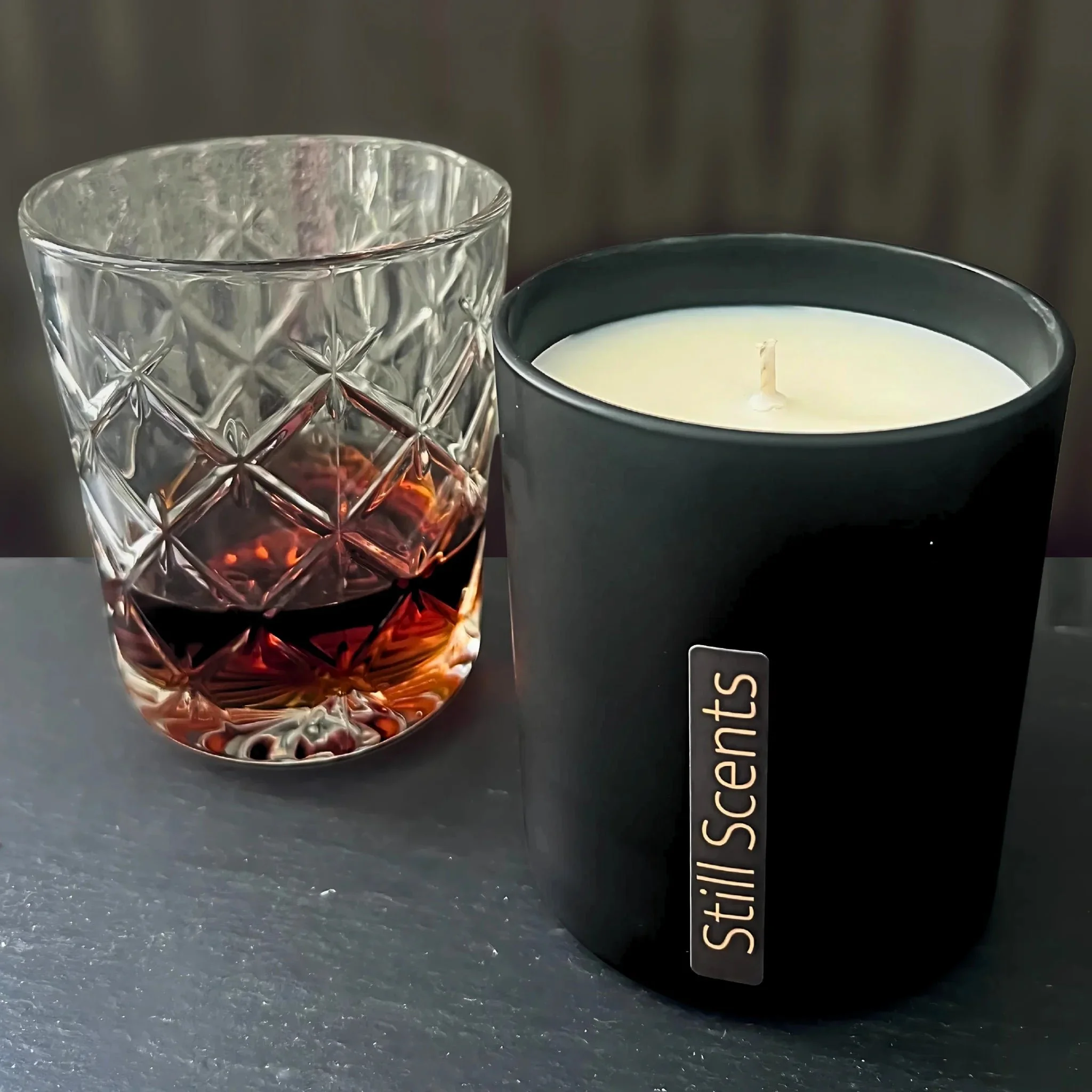 A crystal glass of whisky and a black candle holder with a white lit candle on a dark surface.