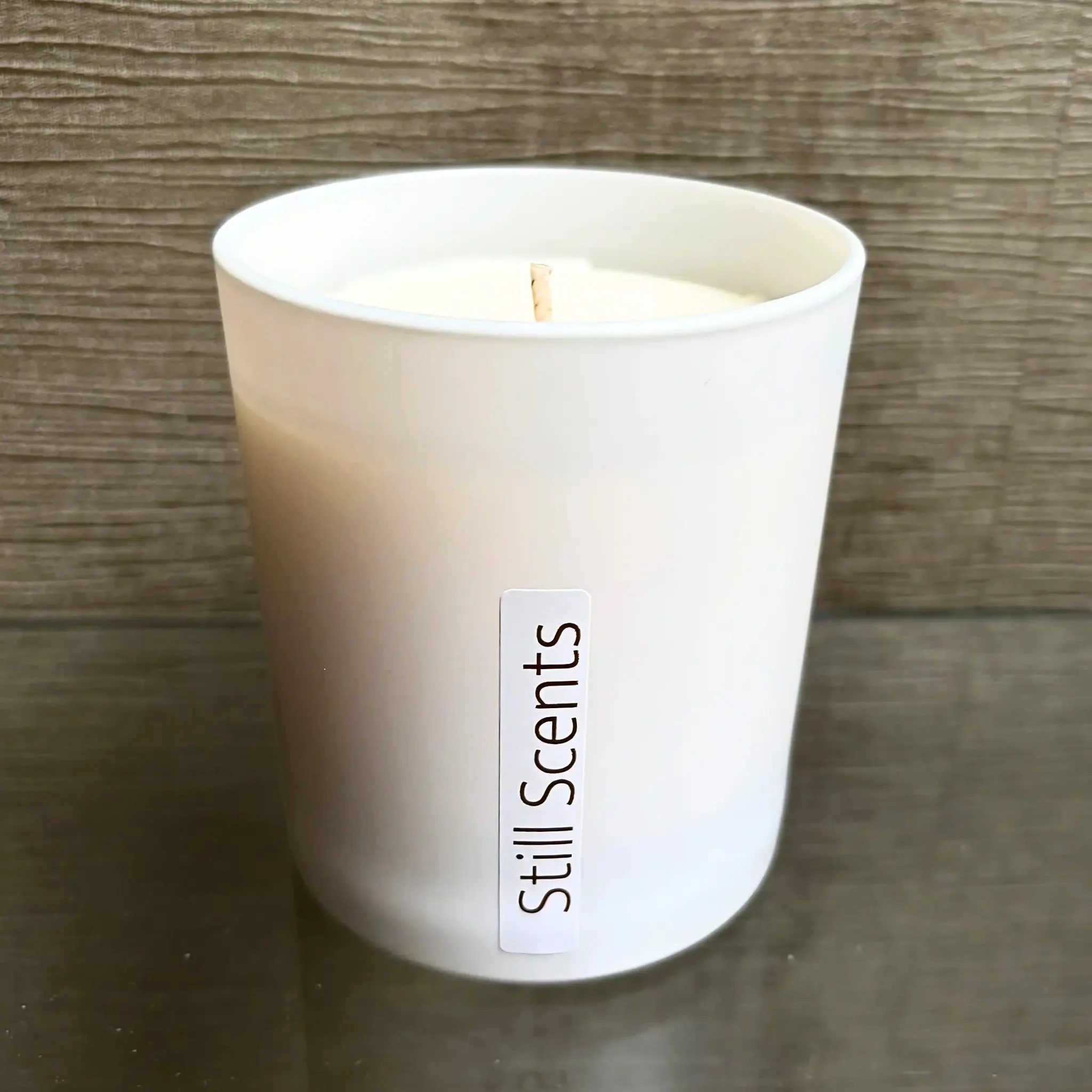 A white candle in a white container with a label reading 'Still Scents' against a wooden background.