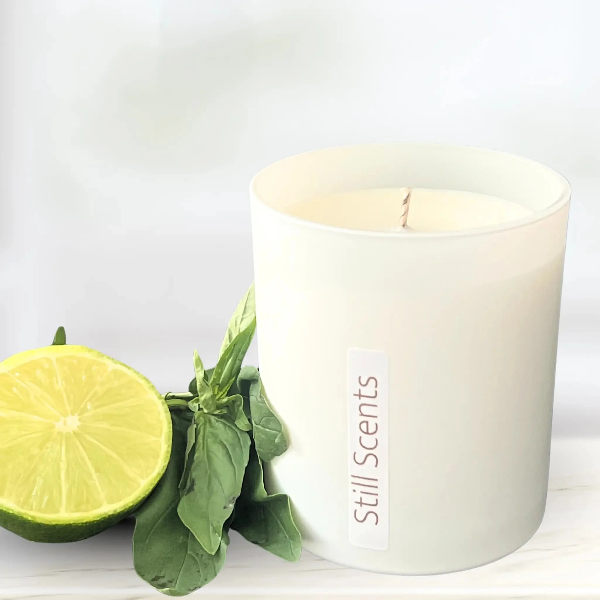 A white candle in a glass jar labeled 'Still Scents' next to a halved lemon and a sprig of fresh basil.