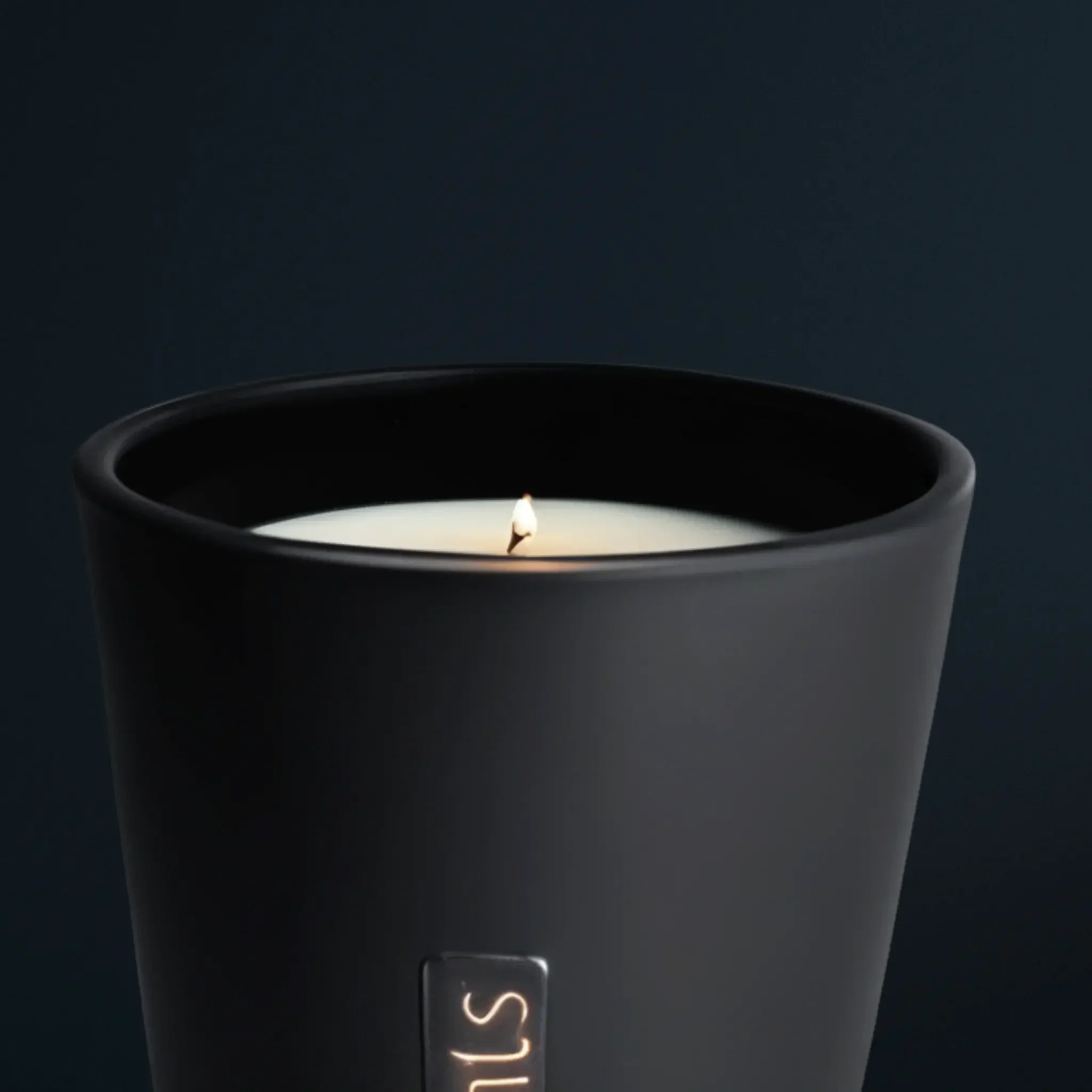 A close-up of a black candle holder with a burning white candle inside, against a dark background.