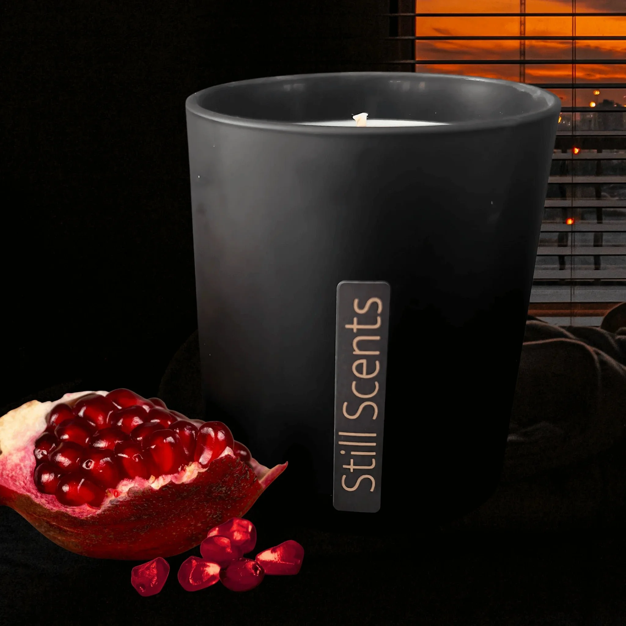 A black candle holder with a candle inside, labeled 'Still Scenes,' along with a halved pomegranate with seeds spilling out, set against a dark background with window blinds and a sunset visible outside.