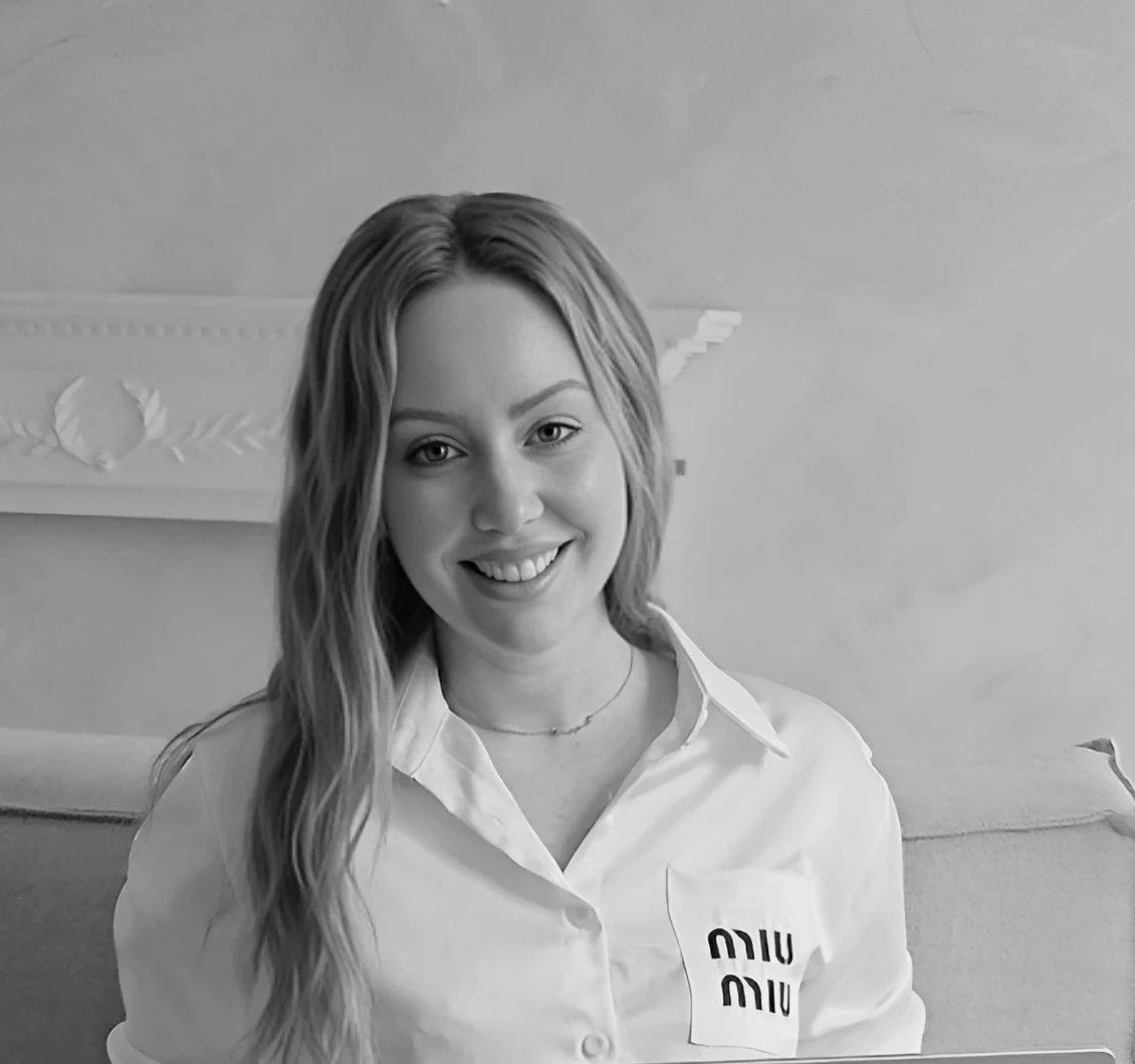 A smiling young woman with long wavy hair, wearing a white shirt with 'miu miu' logo, sitting indoors in front of a decorative wall.