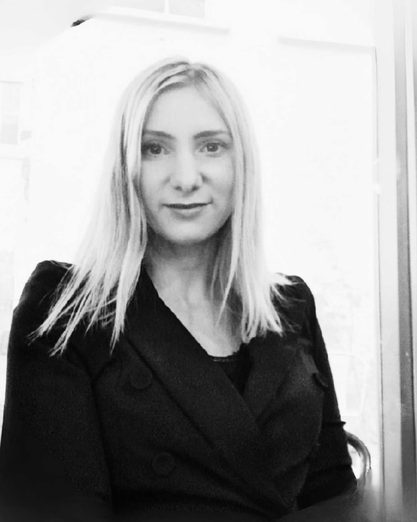 A black and white photo of a woman with long blonde hair, wearing a dark blazer, sitting with her arms folded over her lap, in an indoor setting.