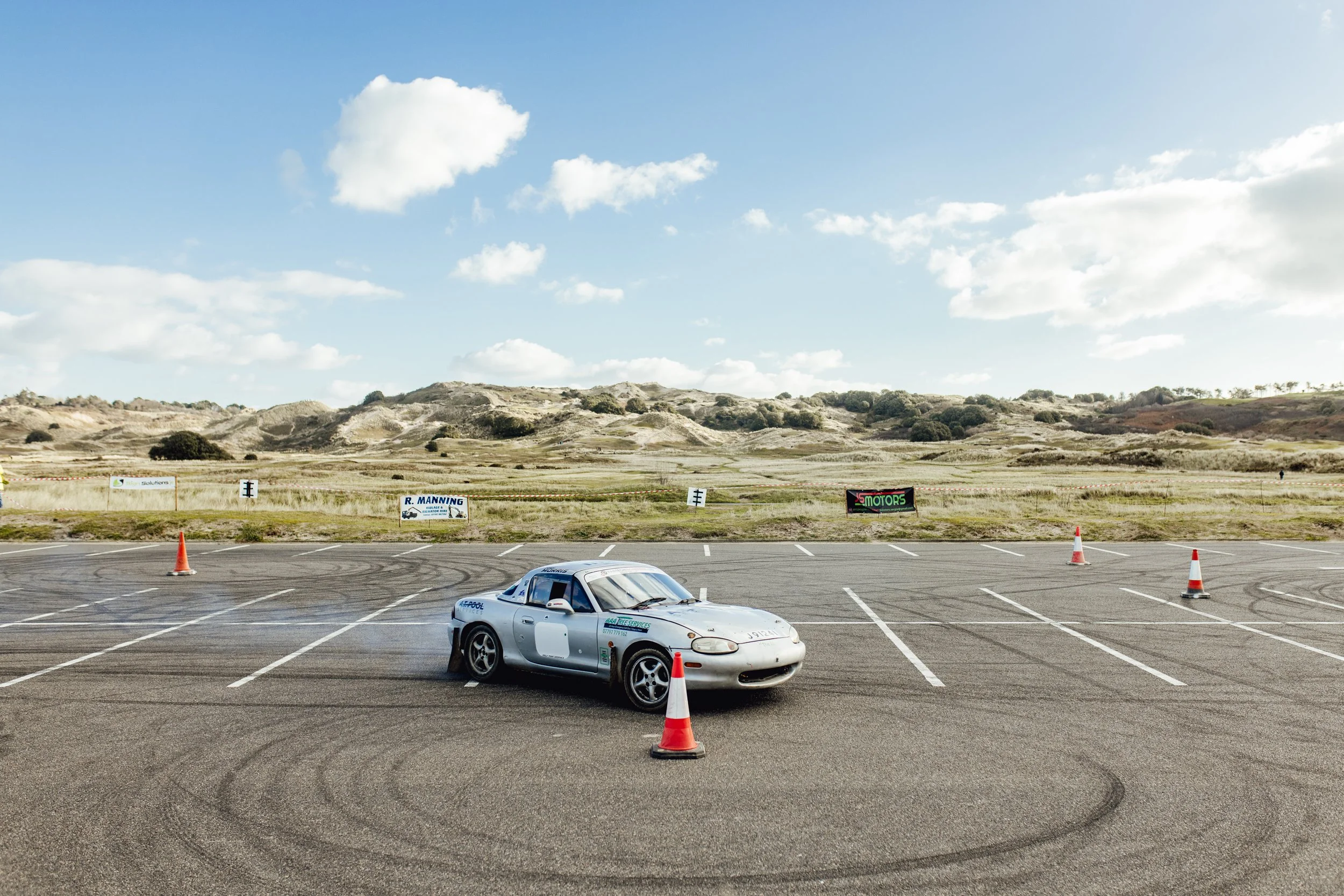 Silver race car on a drifting course with orange cones, tire marks on the asphalt, barren landscape with hills, and a few banners in the background, under a partly cloudy sky.