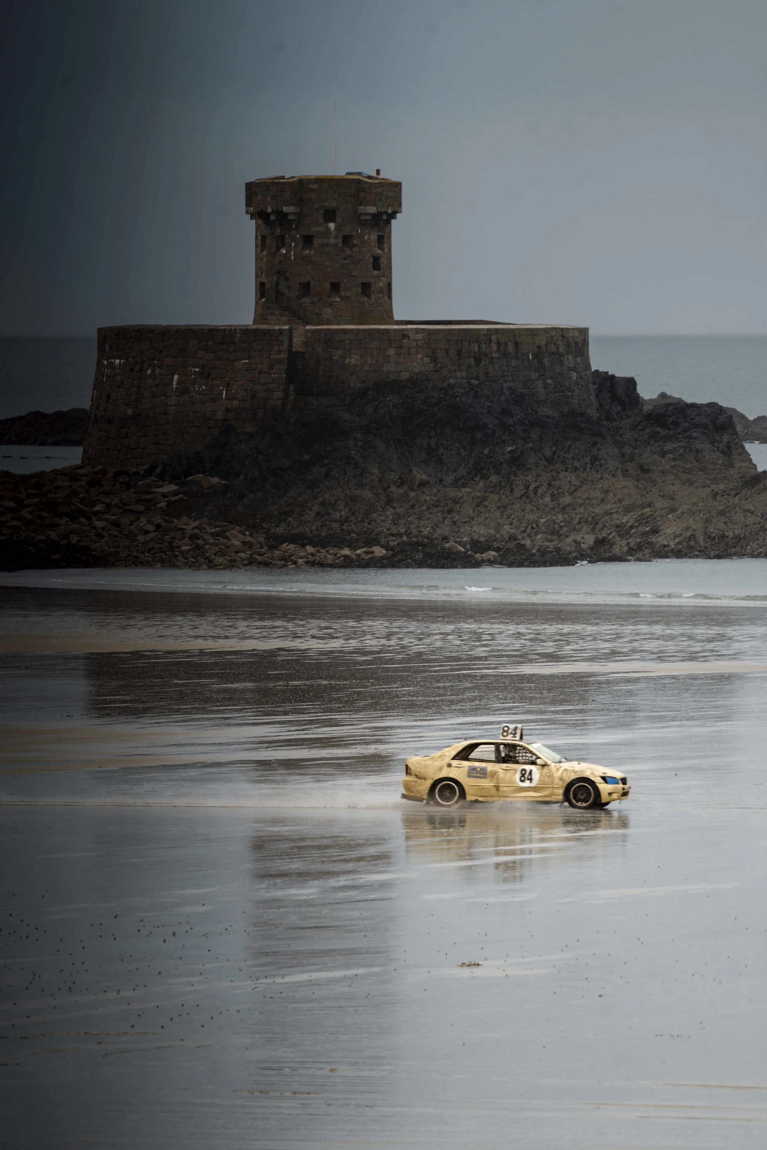 A weathered yellow race car with the number 84 on its side drives along a wet beach with reflections, with a historic stone fortress on a rocky outcrop in the background.