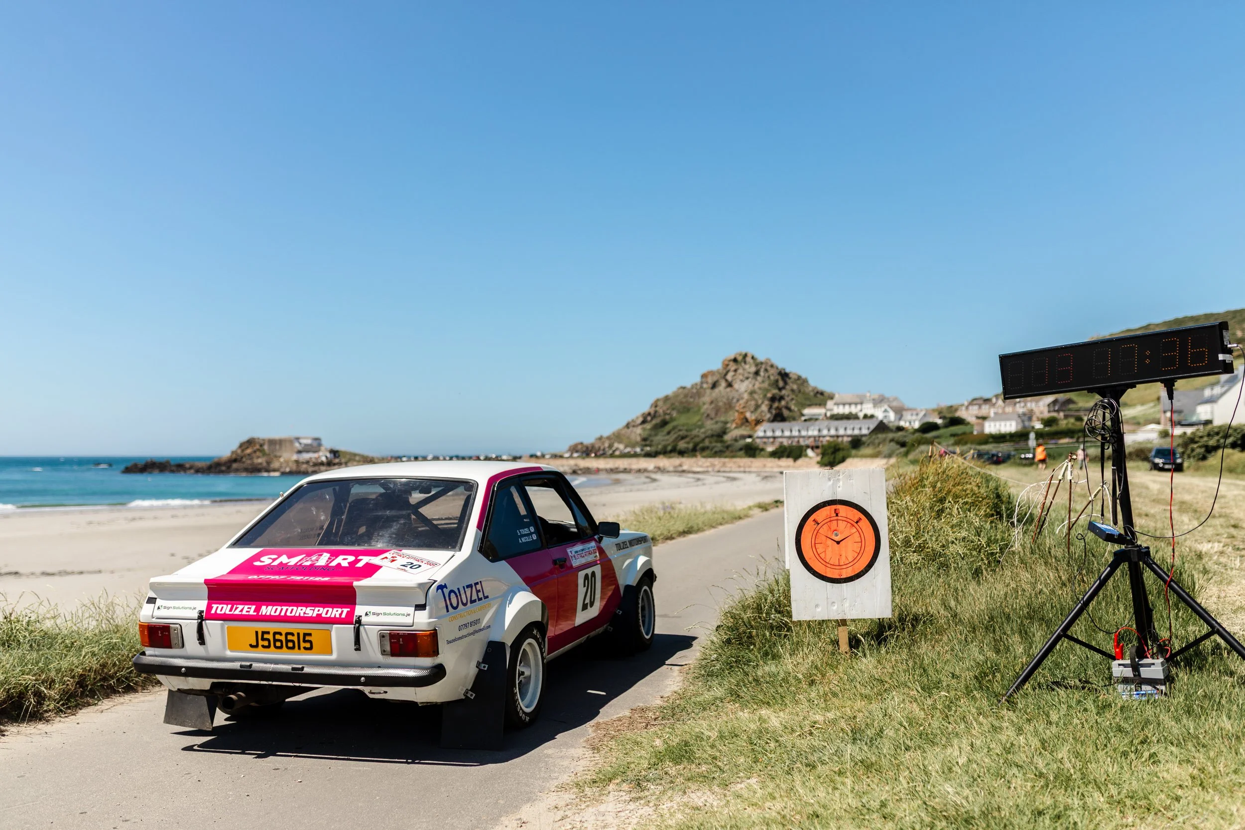 A white race car with pink and blue decals on a seaside road, near a coastal town with hills and buildings in the background, during a motorsport event.