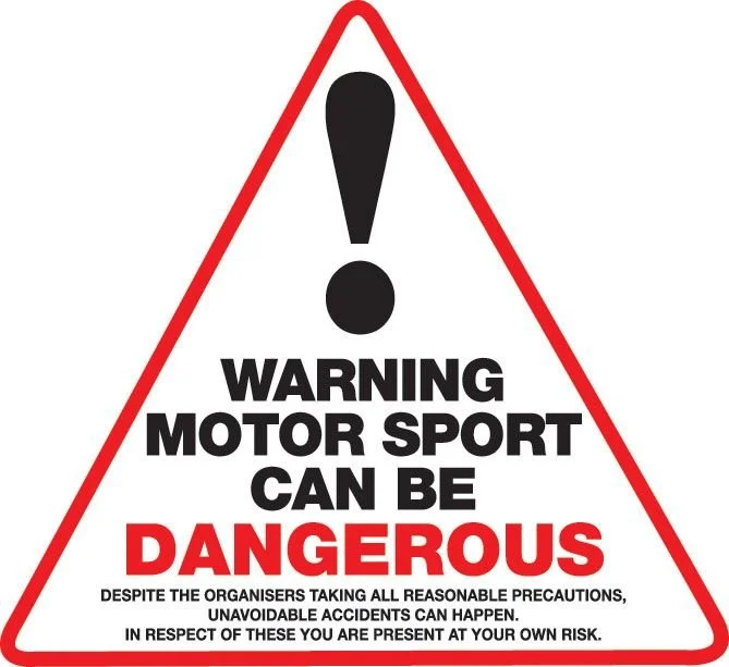 Triangular warning sign with black exclamation mark and bold text warning about motor sport dangers, advising to take precautions and acknowledge the risk.