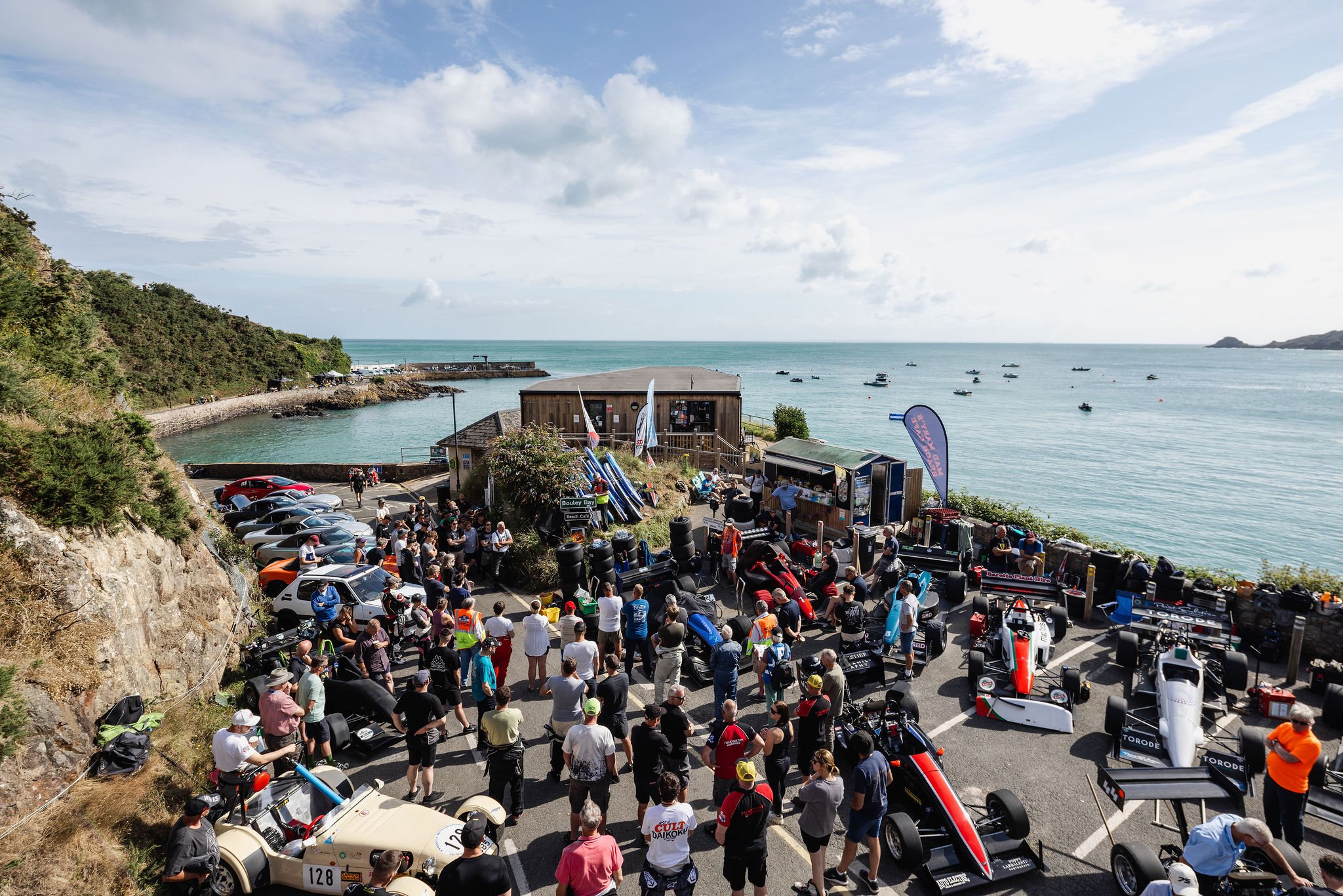 Crowd of people and race cars in a parking lot near the ocean, with some cars and boats in the water, and a wooden building on the seaside.