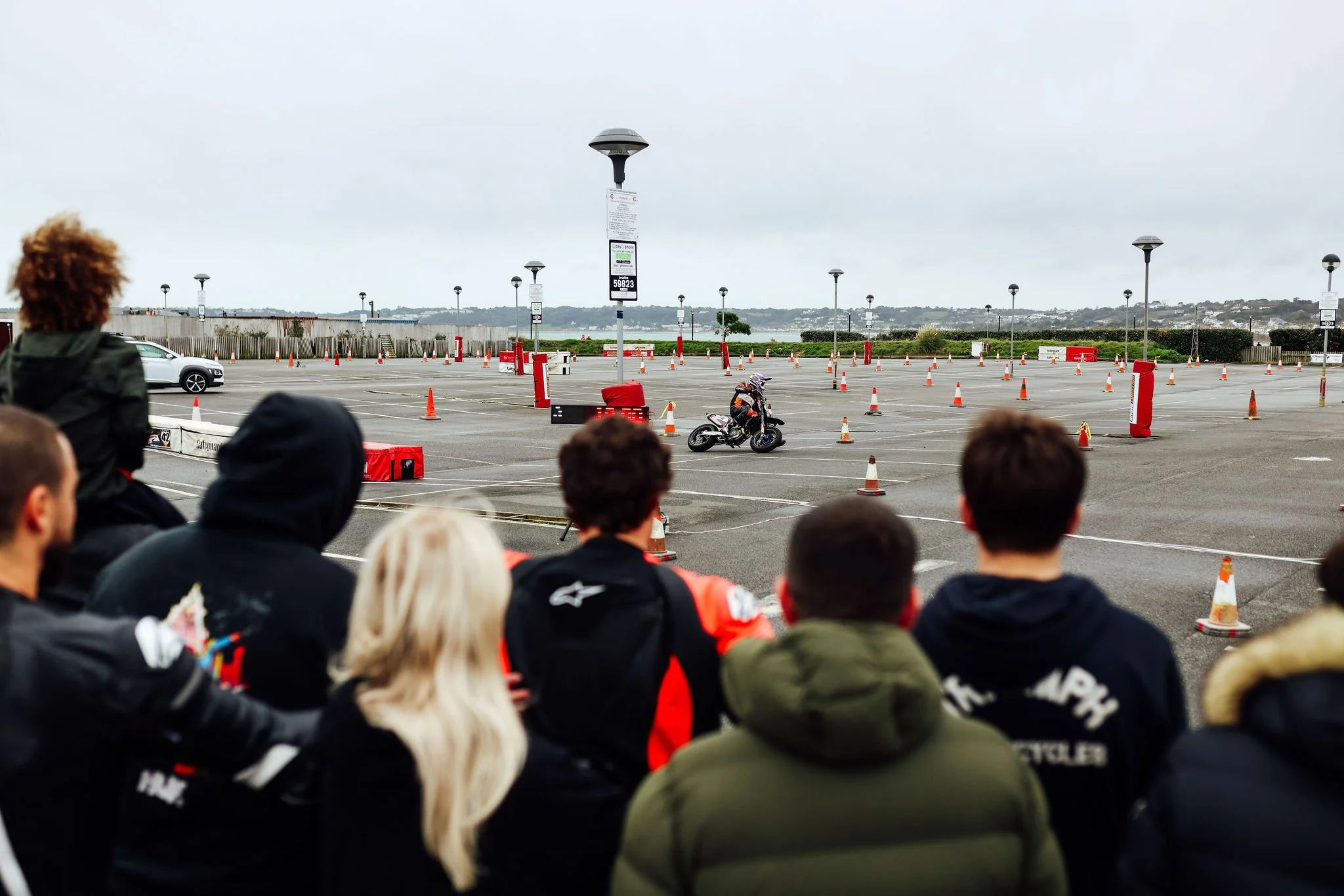 A group of people watching a motorcycle rider practicing on an outdoor parking lot used as a closed course, with traffic cones and barriers, on a cloudy day.