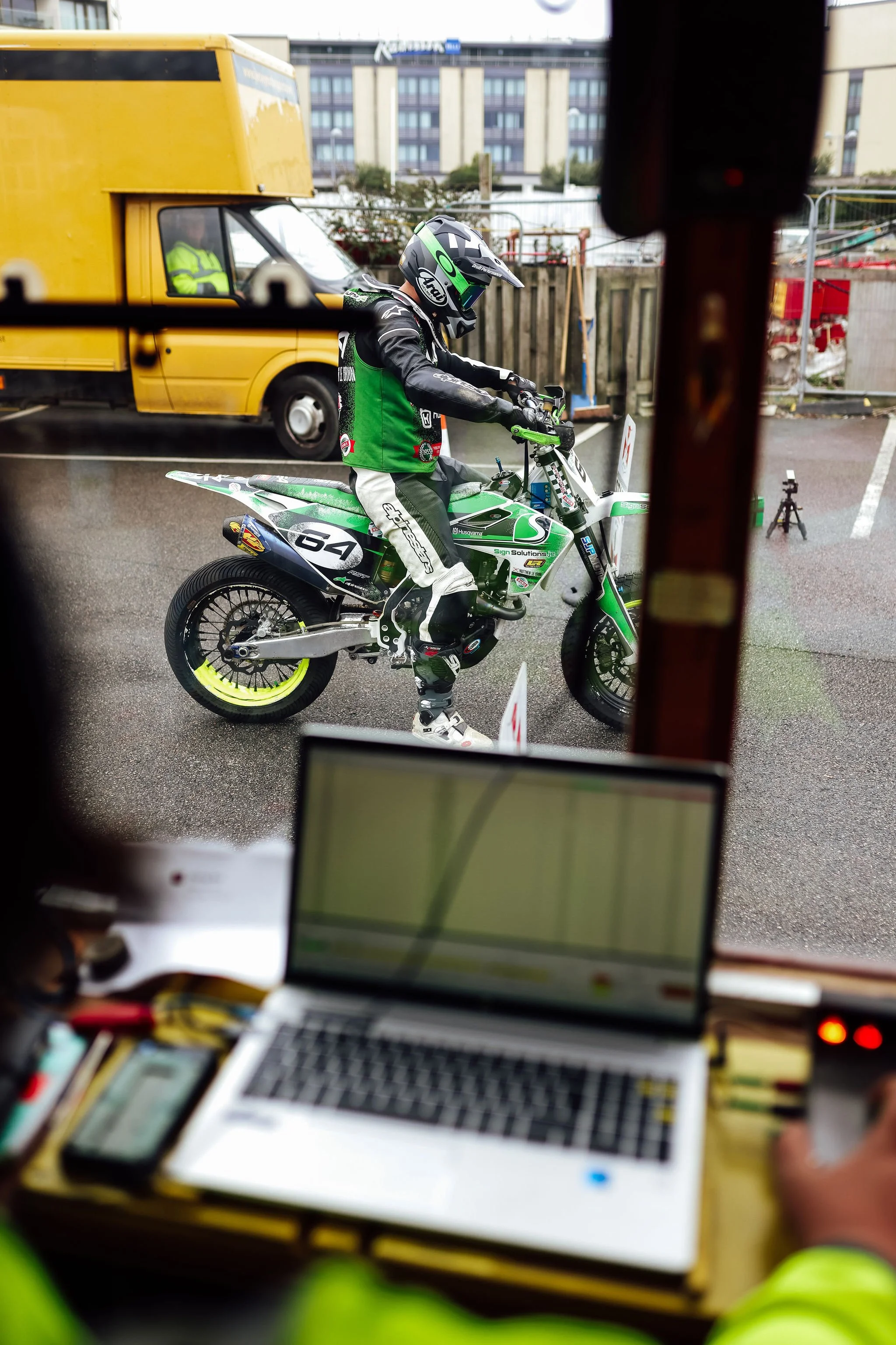 A motorcyclist in racing gear and helmet sitting on a green and white dirt bike outside near a yellow delivery truck and urban buildings.