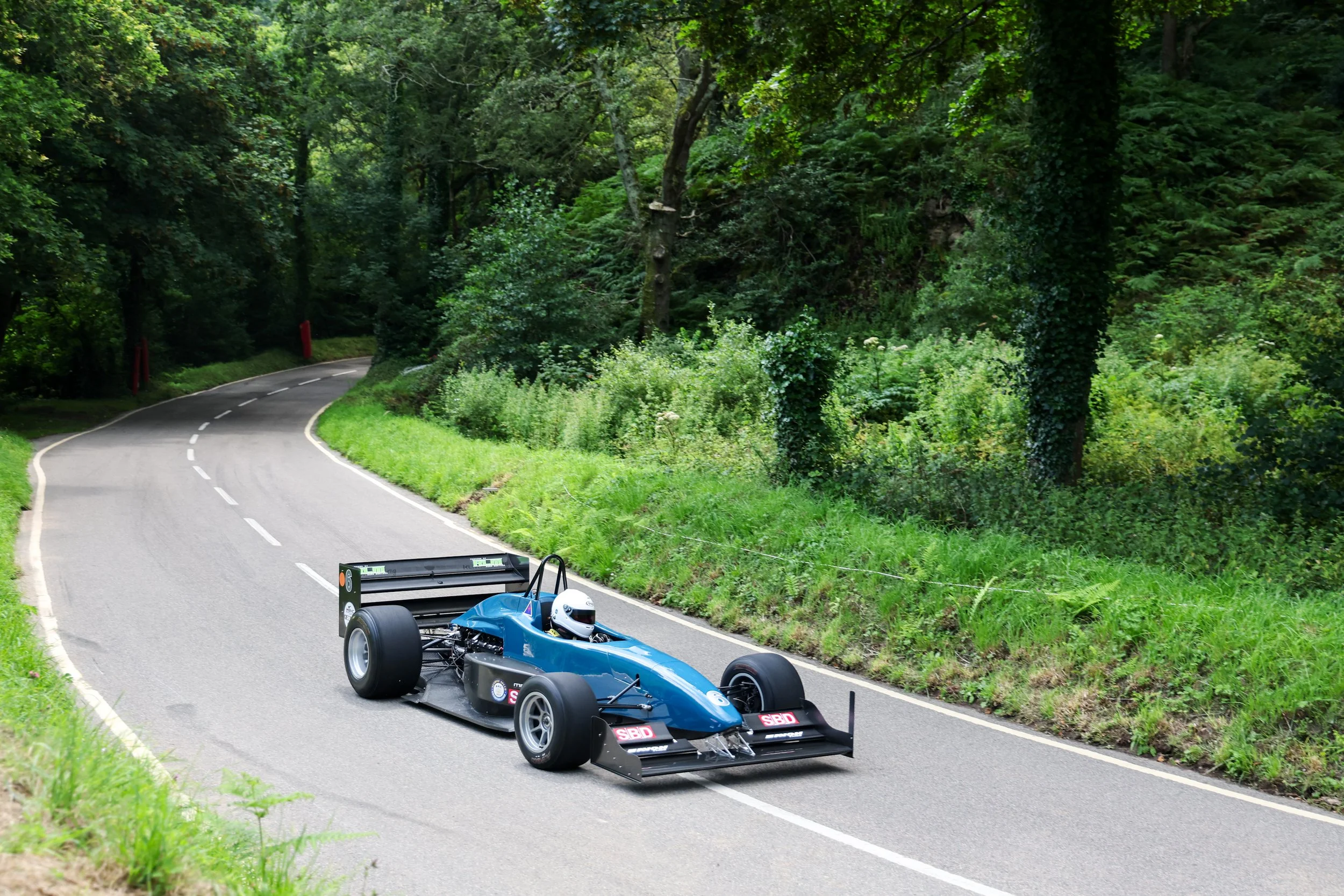 A blue open-wheel race car driving on a winding, green forested road with dense trees and bushes on either side.