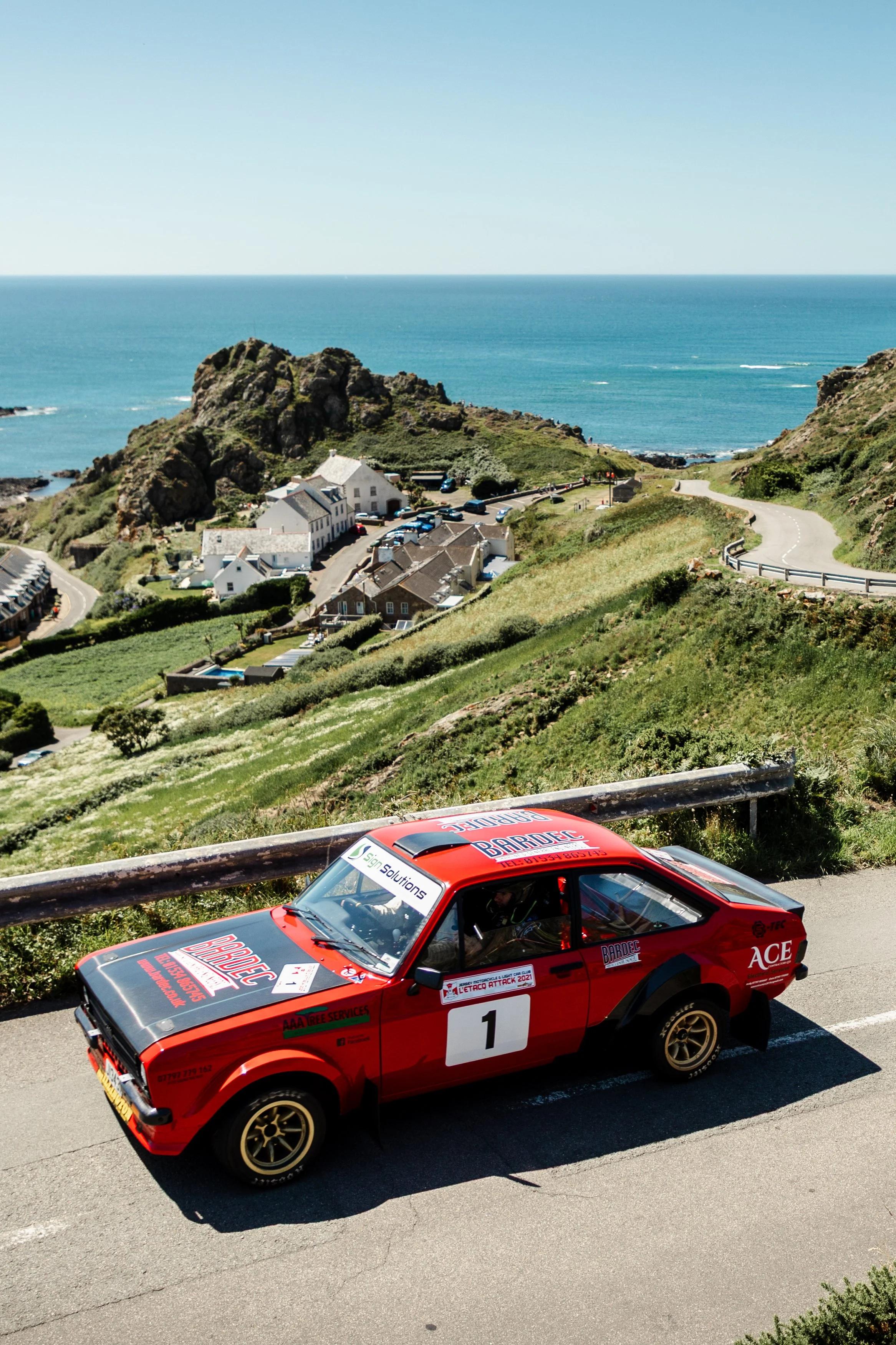 A red vintage rally car with the number 1 on its side drives along a coastal mountain road with a scenic view of a village and the ocean in the background.