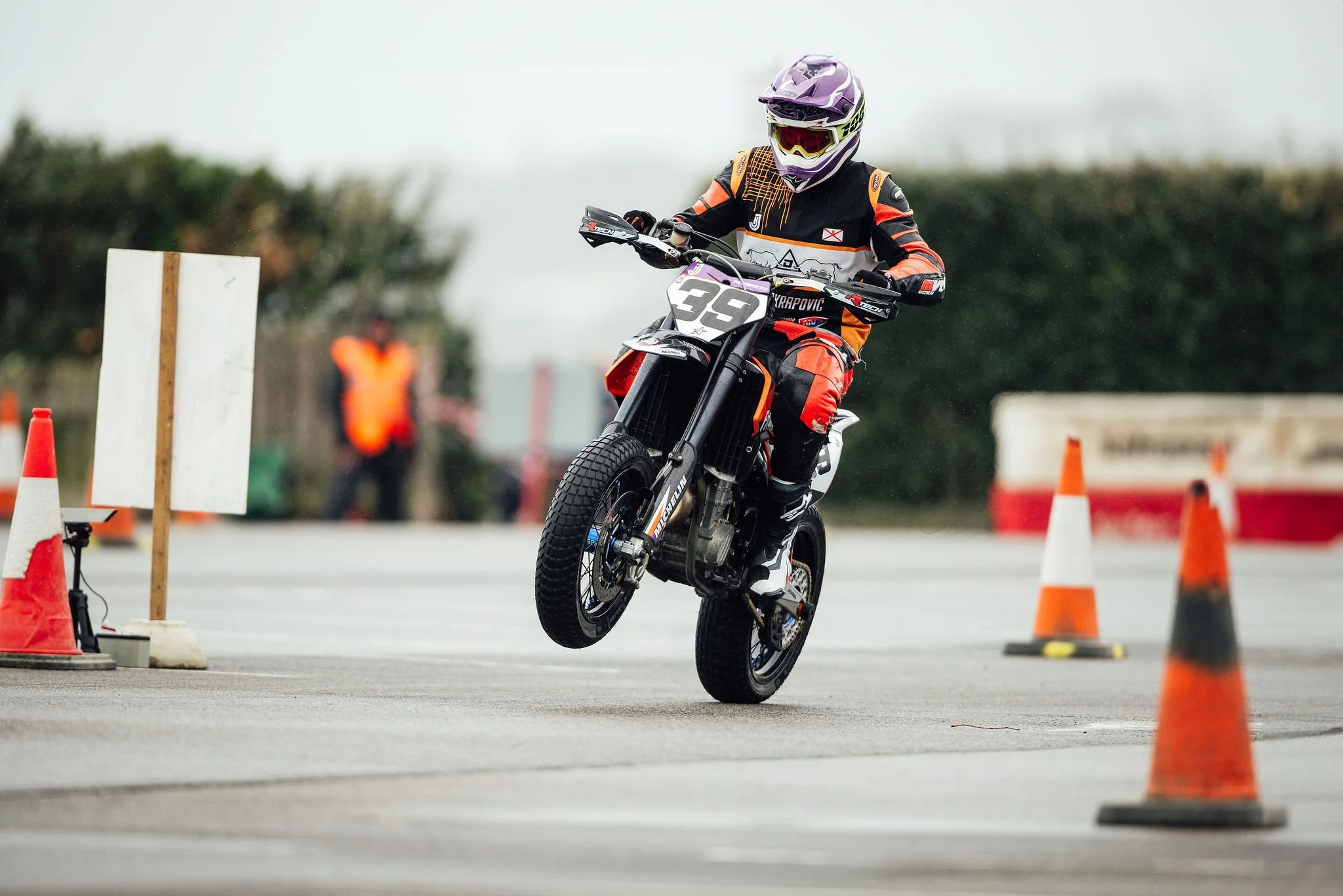 Motocross rider in orange and black gear performing a wheelie on a dirt bike on an outdoor course with orange traffic cones.