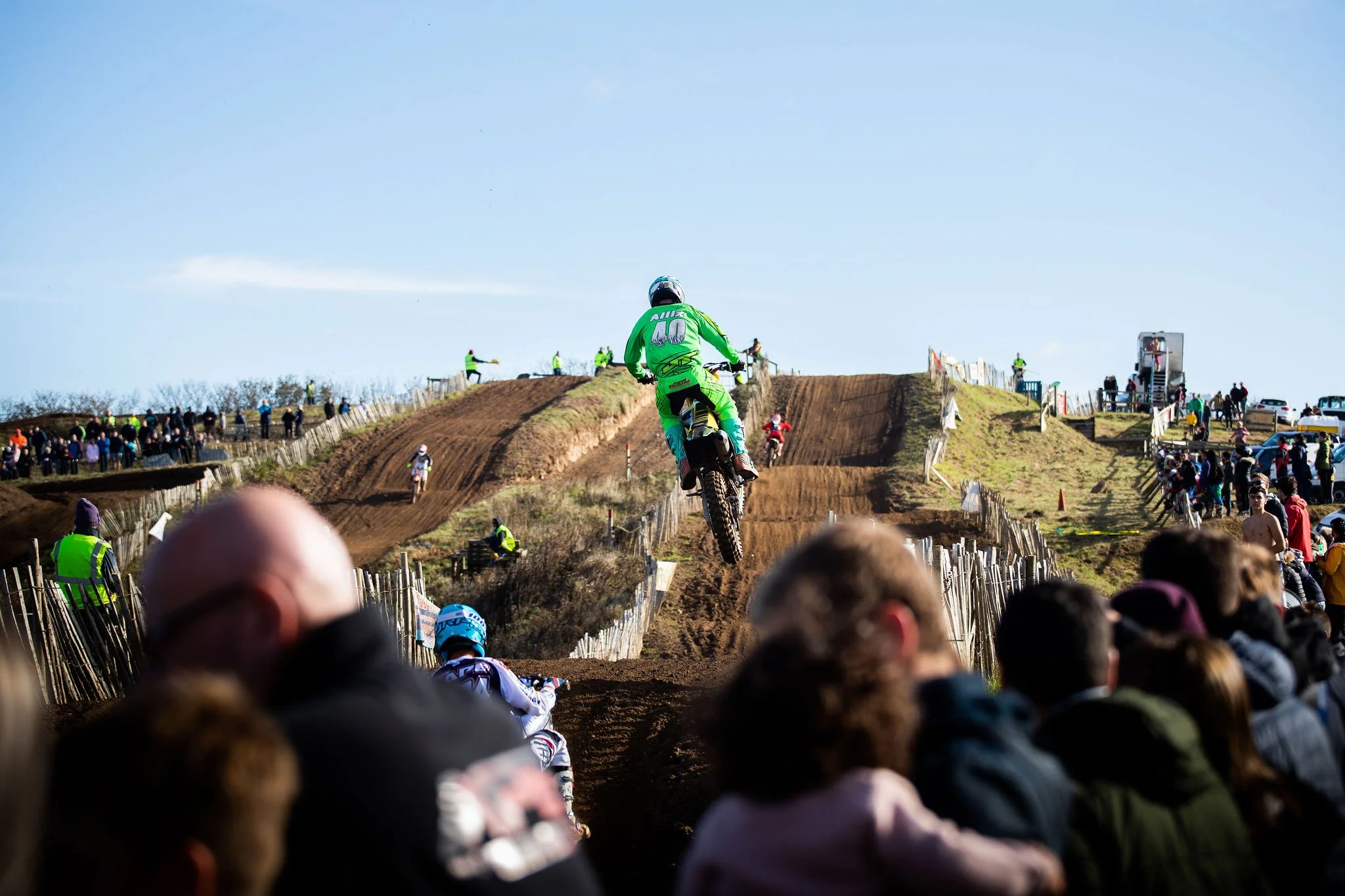 Motorcycle rider in green gear mid-air over a dirt jump during a motocross race, with spectators and officials watching from the sides of the track under a clear sky.