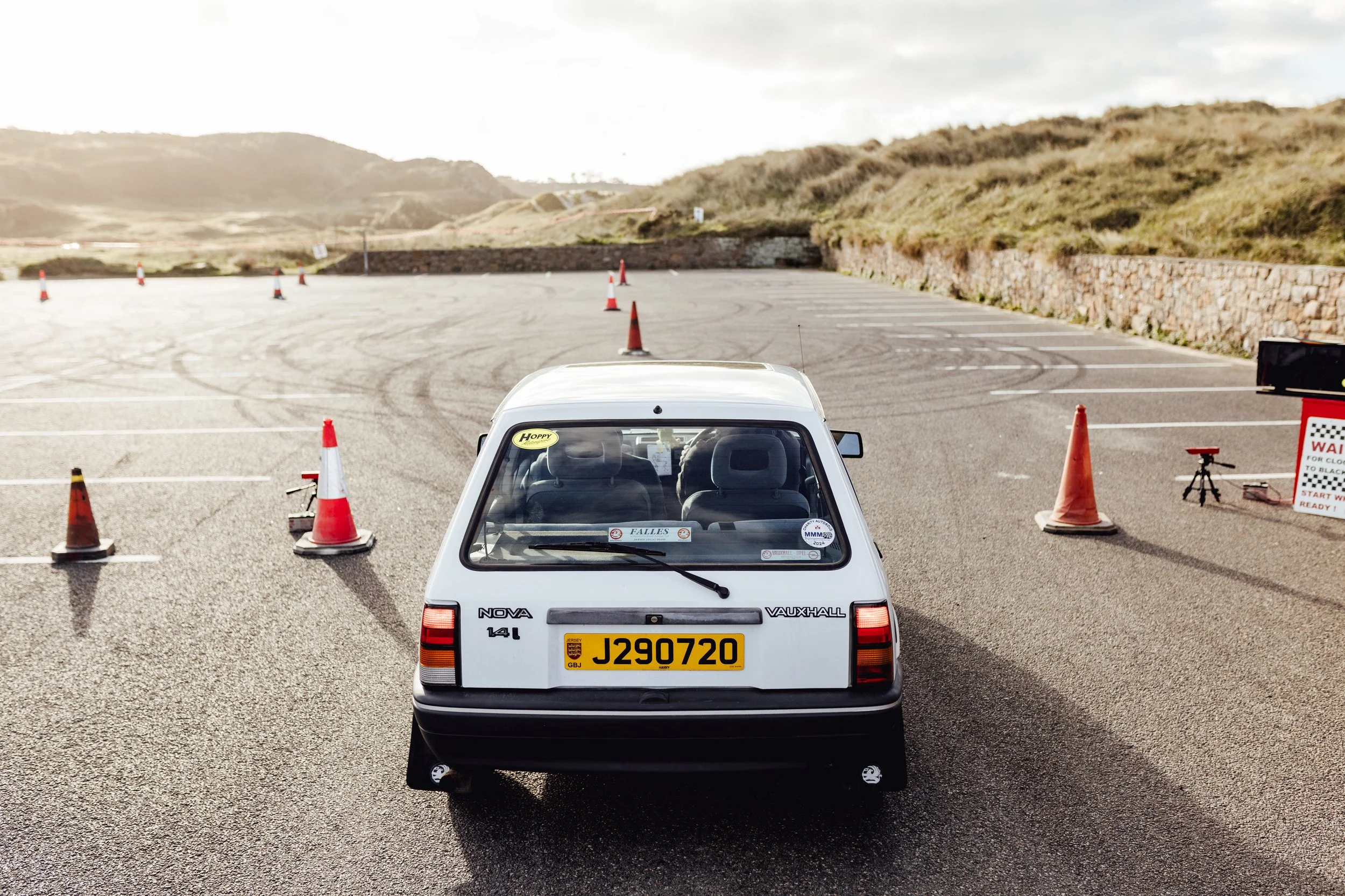 White Vauxhall Nova hatchback parked in an outdoor parking lot with orange traffic cones, rolling hills in the background, and a sign on the right.