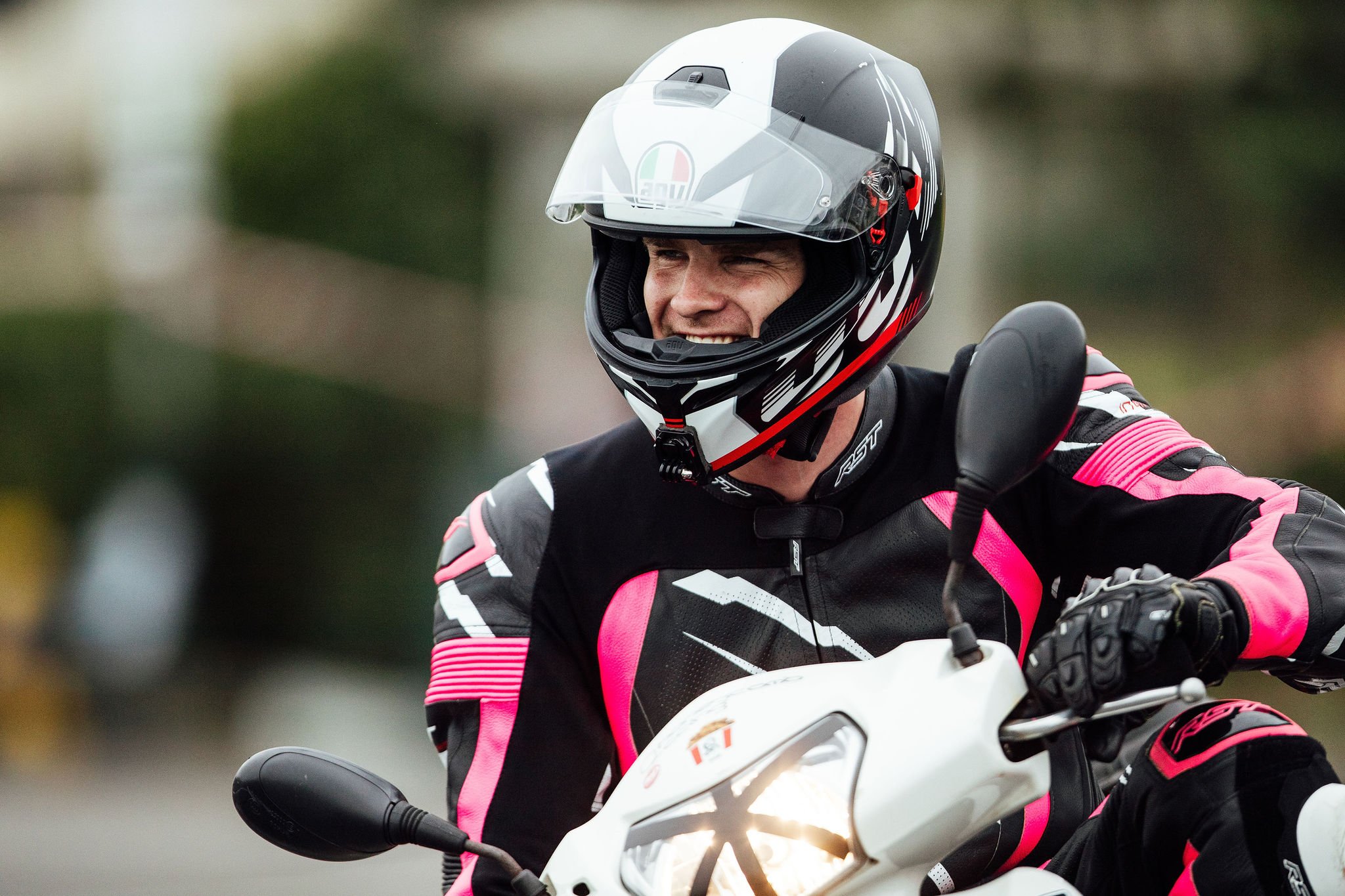 Motorcycle rider wearing a black and pink racing suit and a full-face helmet, smiling while riding a white motorbike.