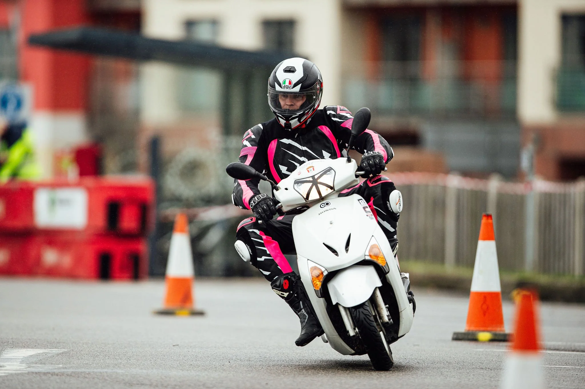 A person wearing a black helmet and riding a white scooter navigates through orange traffic cones on a city street.