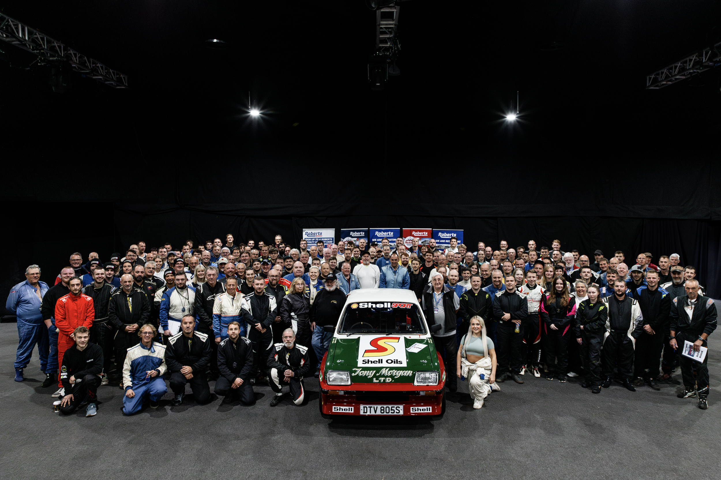 Large group photo of competitors and supporters at the Jersey Rally, posing indoors behind a rally car.